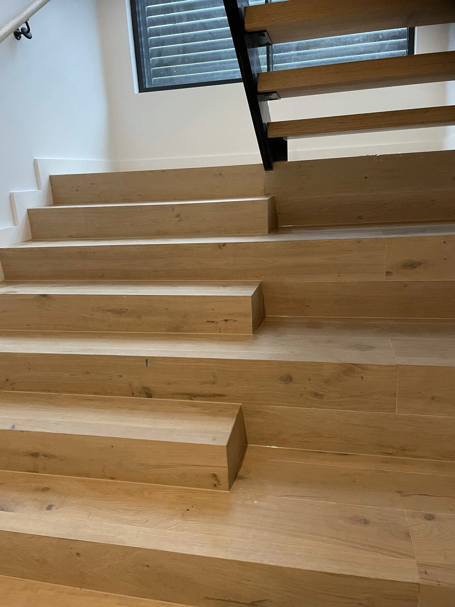 Wooden staircase with a black metal handrail leading up near a window with black shutters.