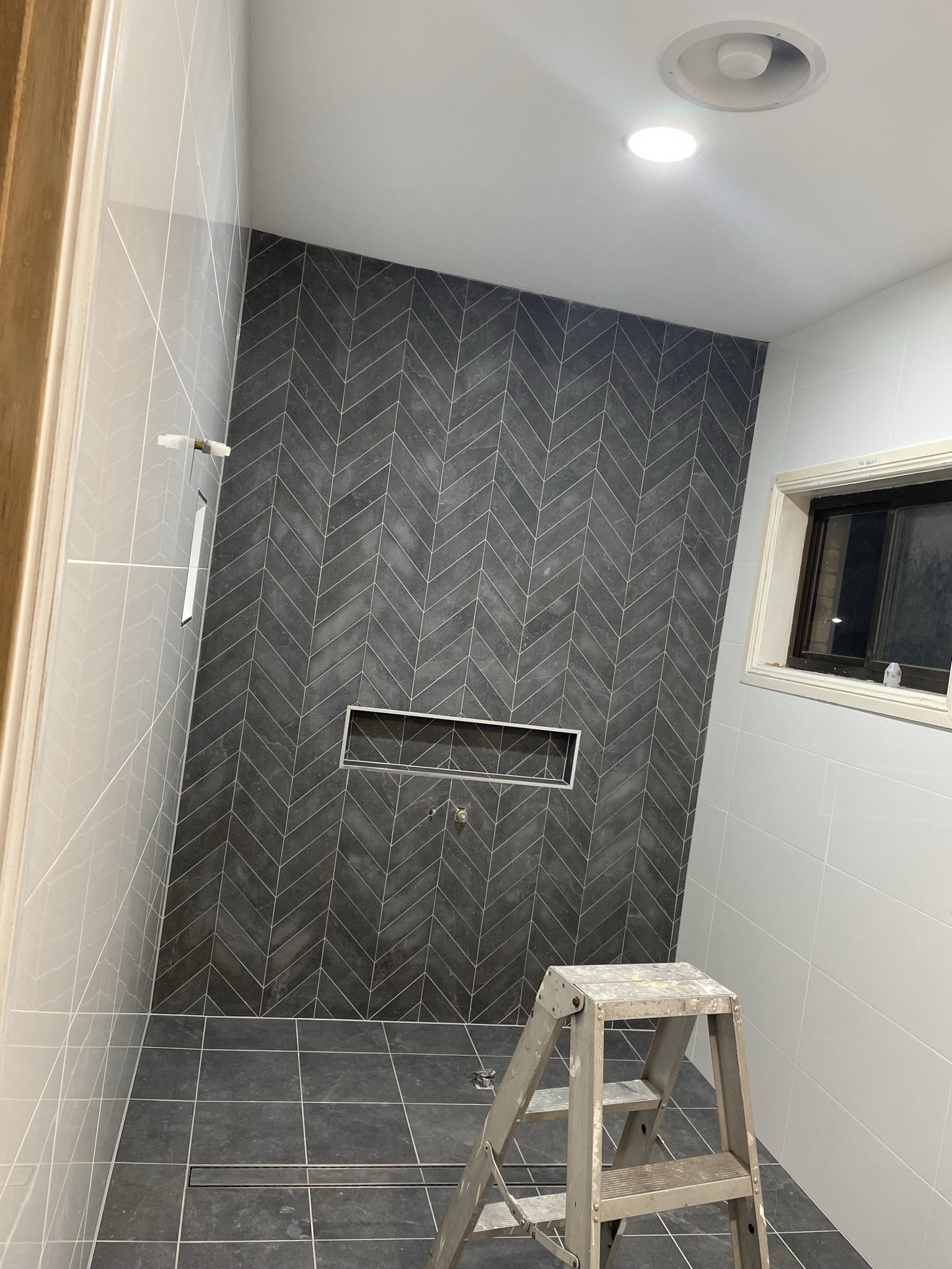Bathroom under construction with gray herringbone tiled wall, white tiled side wall, small window, ceiling light, and a stepladder