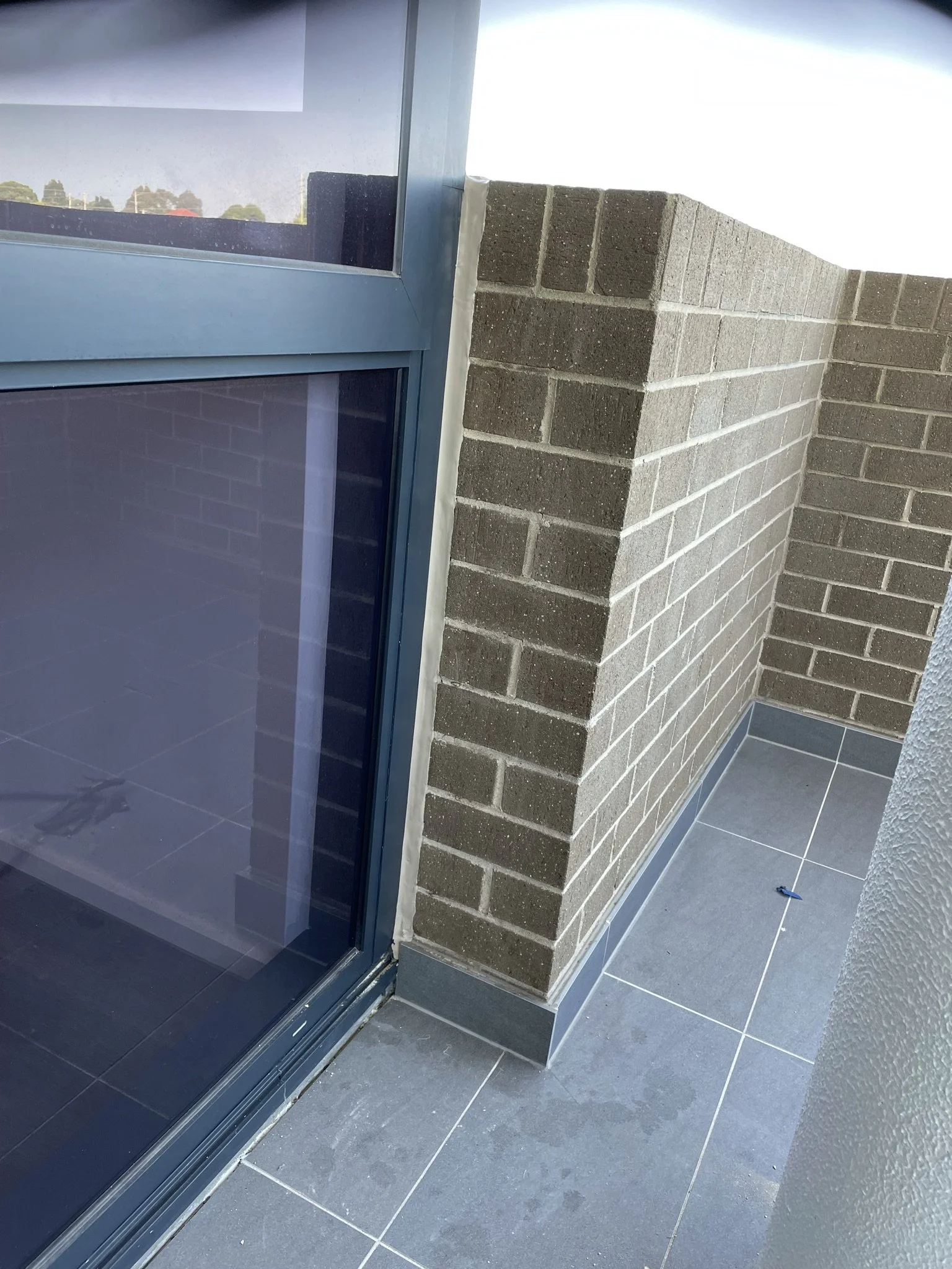 A corner of a balcony or patio with gray tiled flooring, a brick wall, and a door with glass panes and a blue frame. The sky is visible in the background.