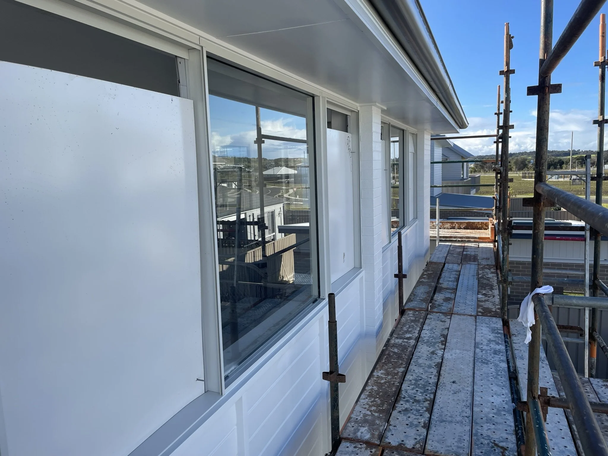 Construction scaffolding on the side of a white building with large windows, overlooking a suburban landscape on a sunny day.