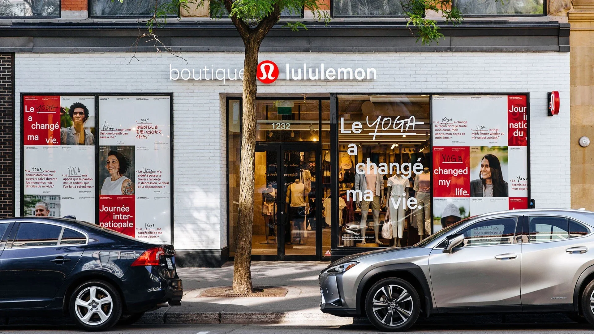 Exterior of a lululemon store with mannequins and clothing inside, large window posters with images of people and text in French and English, parked cars in front, and a tree blocking part of the store sign.