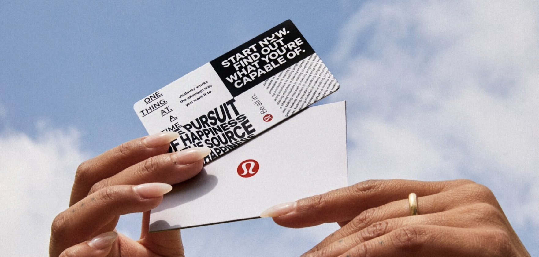 Close-up of a person holding a business card with the Lululemon logo, with a blue sky in the background.