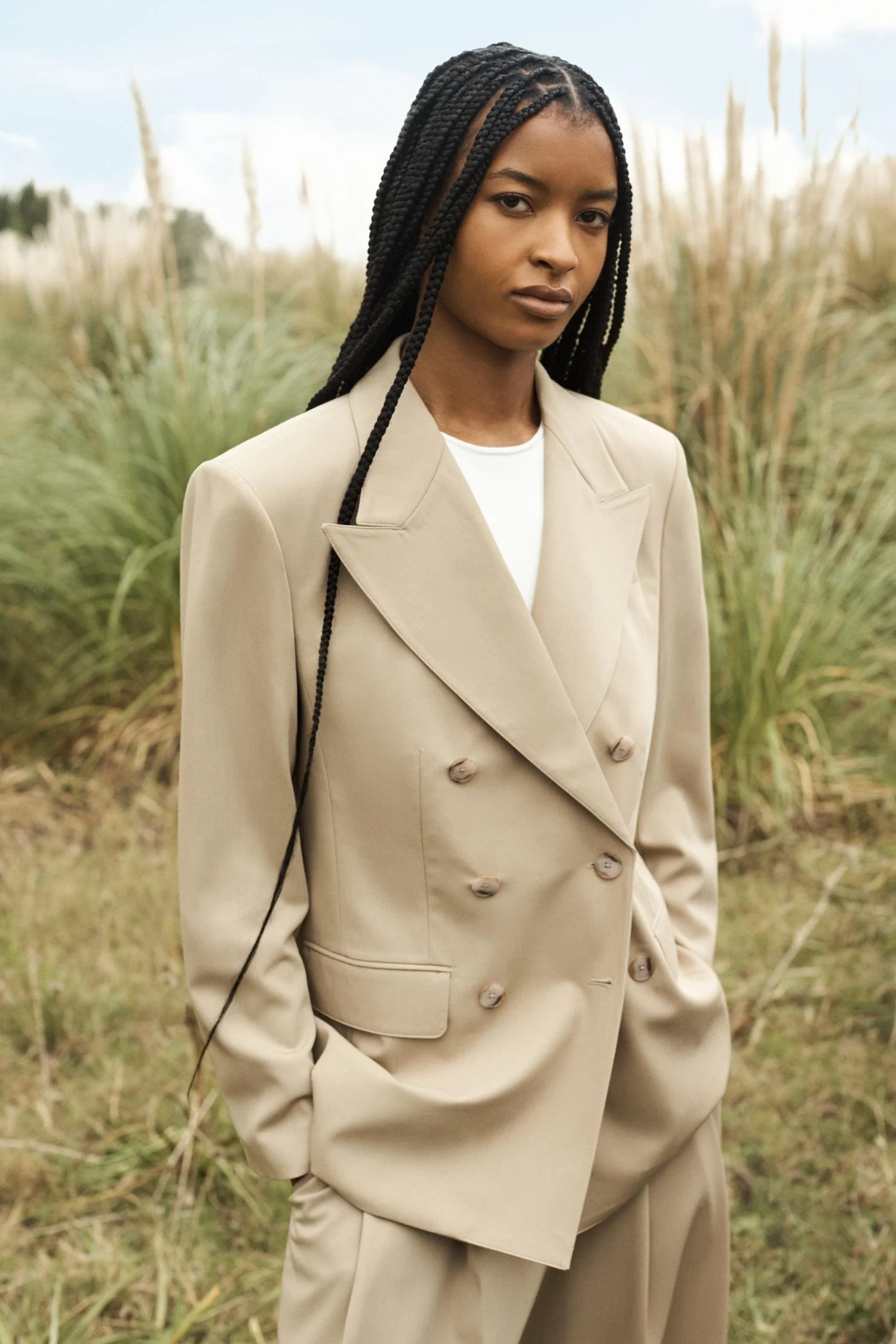 A woman in a beige oversized double-breasted suit stands outdoors in a grassy field, with her hands in her pockets, looking at the camera.
