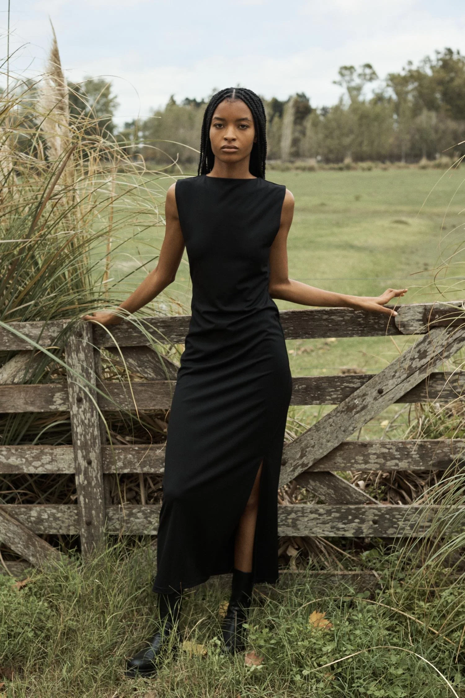A woman in a black sleeveless dress standing outdoors near a weathered wooden fence, with tall grass and trees in the background.
