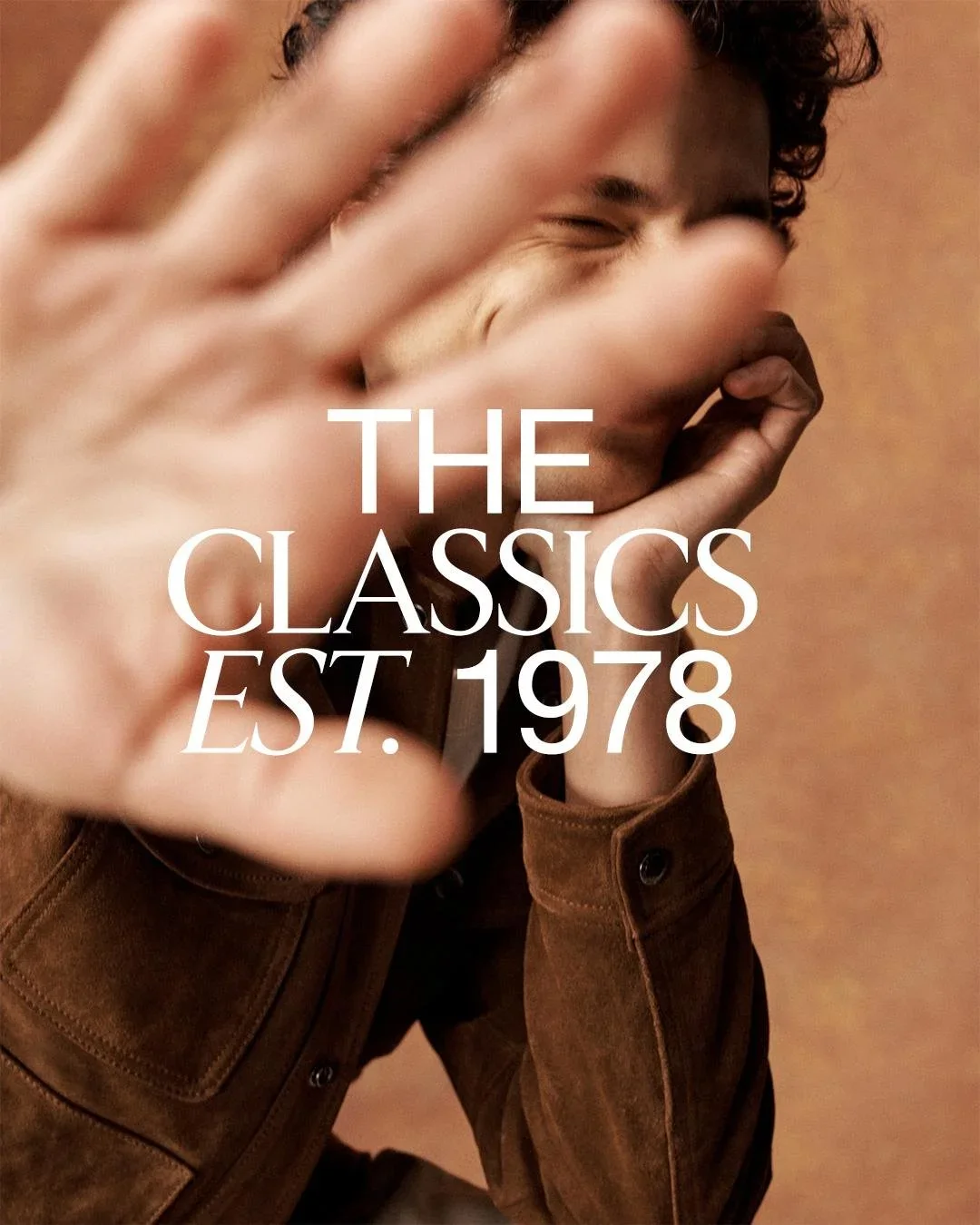 A smiling person with curly hair covers part of their face with their hand, wearing a brown jacket, with text overlay reading 'The Classics Est. 1978'.