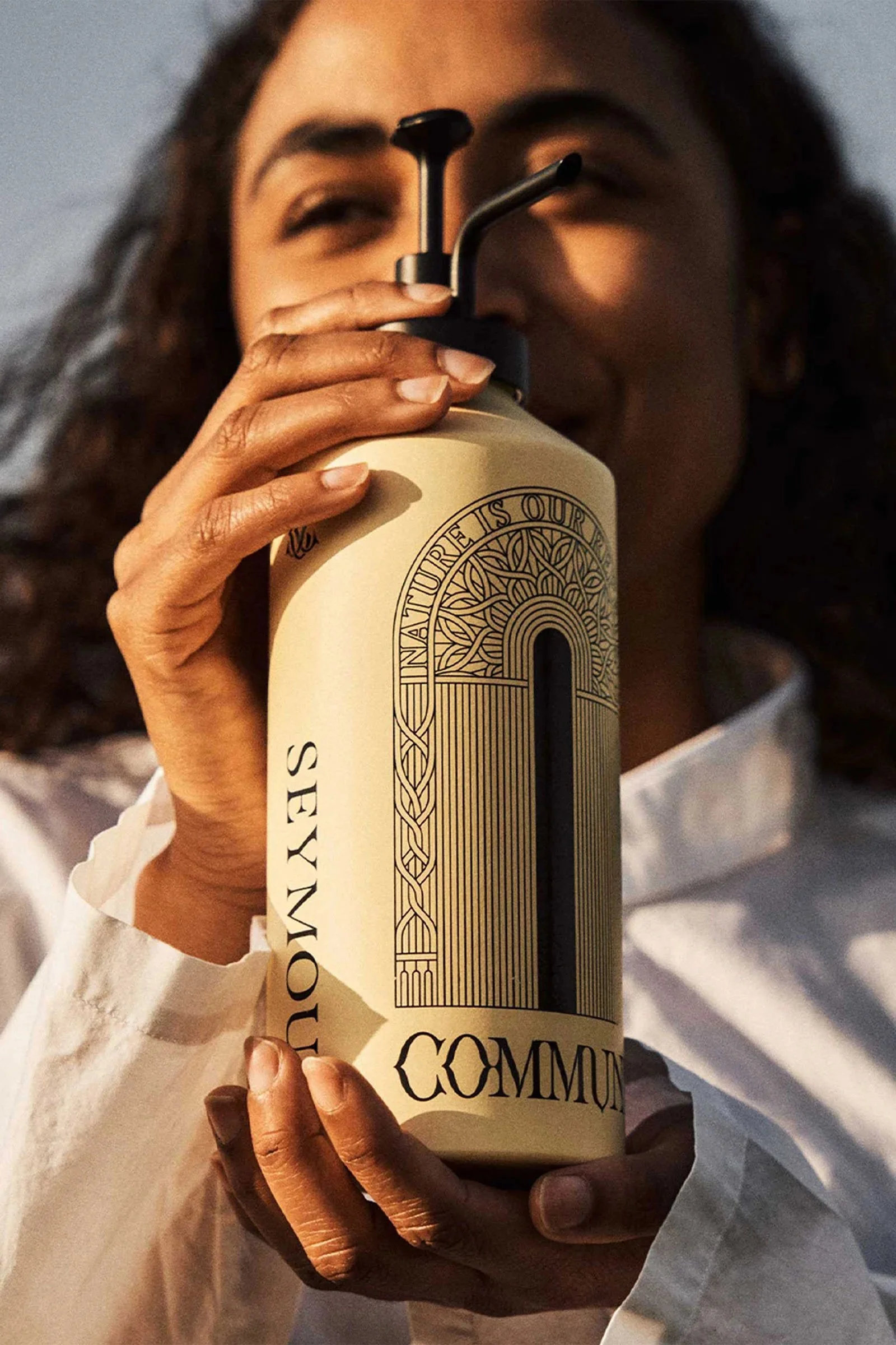 Close-up of a person with medium skin tone holding a beige reusable water bottle with black and beige design and the words 'NATURAL RES OUR' visible. The person has long curly hair and is wearing a white shirt.