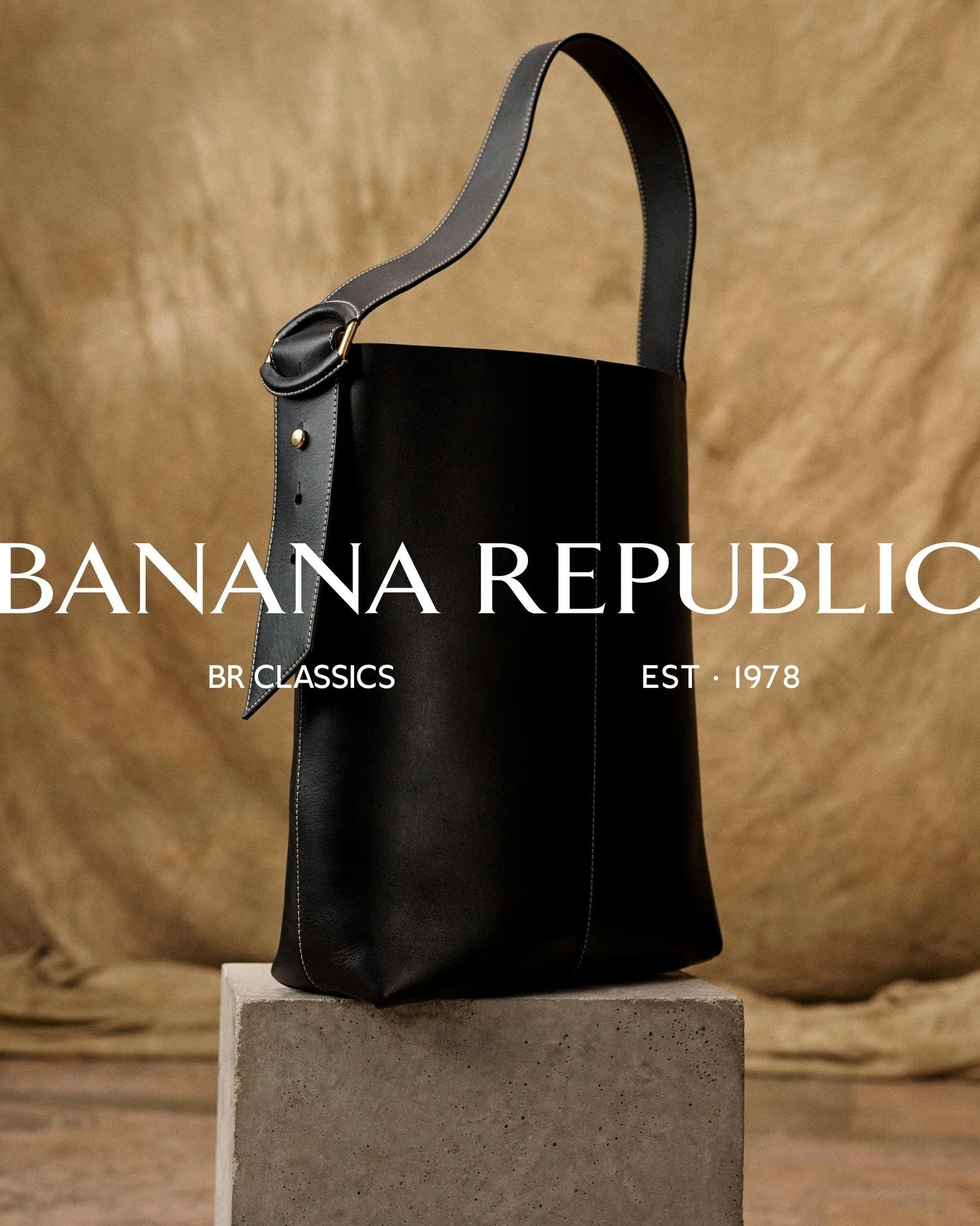 Black leather tote bag with a shoulder strap, displayed on a concrete pedestal against a brown textured background with white text reading 'BANANA REPUBLIC BR CLASSICS EST. 1978'.