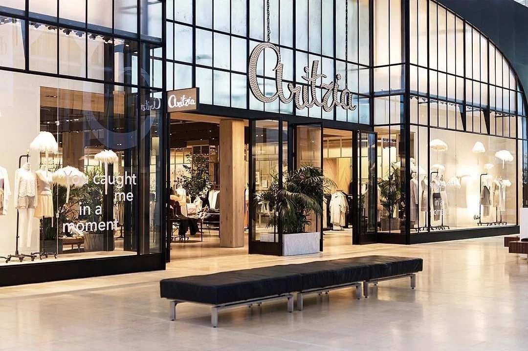 Exterior view of a clothing store called 'Aritzia' in a mall, with glass walls displaying mannequins dressed in women's clothes and indoor plants visible through the windows.