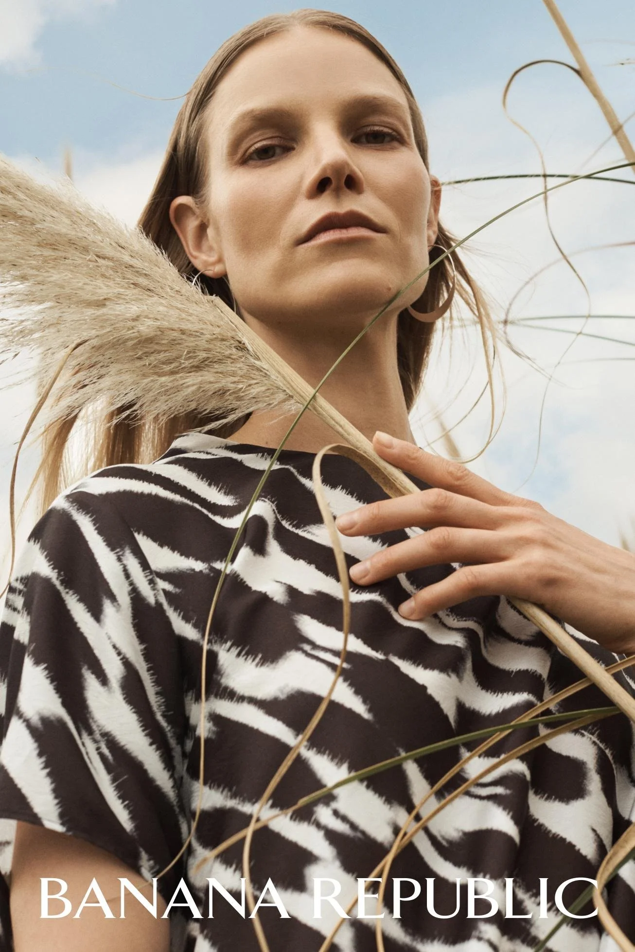 A woman with long hair in natural makeup, wearing a zebra-patterned shirt, holding a large dried grass stalk near her shoulder, outdoors with a cloudy sky background.
