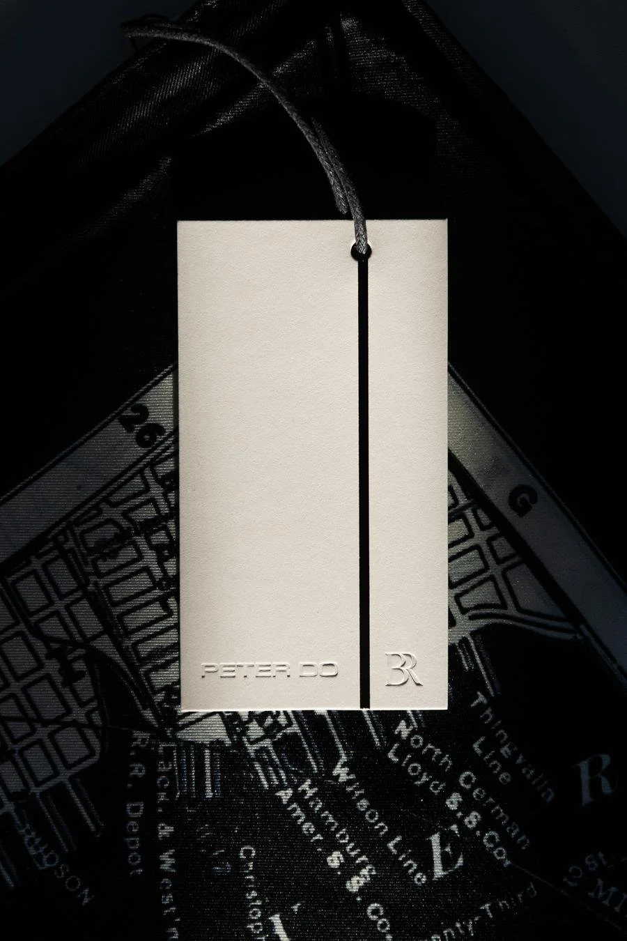 A white tag with black text reading 'PETER DO' and a small logo, attached to a dark fabric garment.