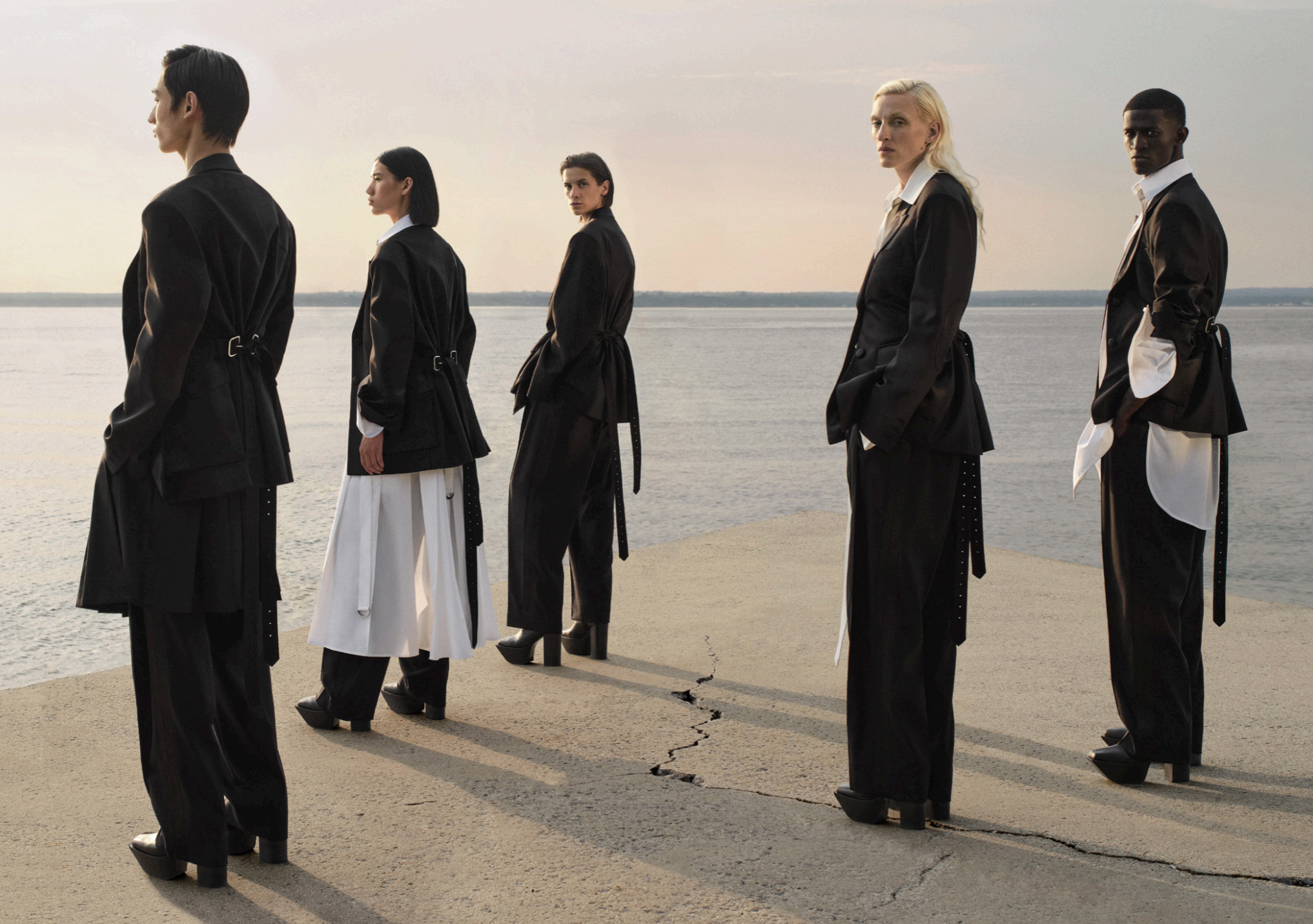 Five models dressed in black and white fashion standing in a line on a cracked concrete surface by a body of water at sunset.