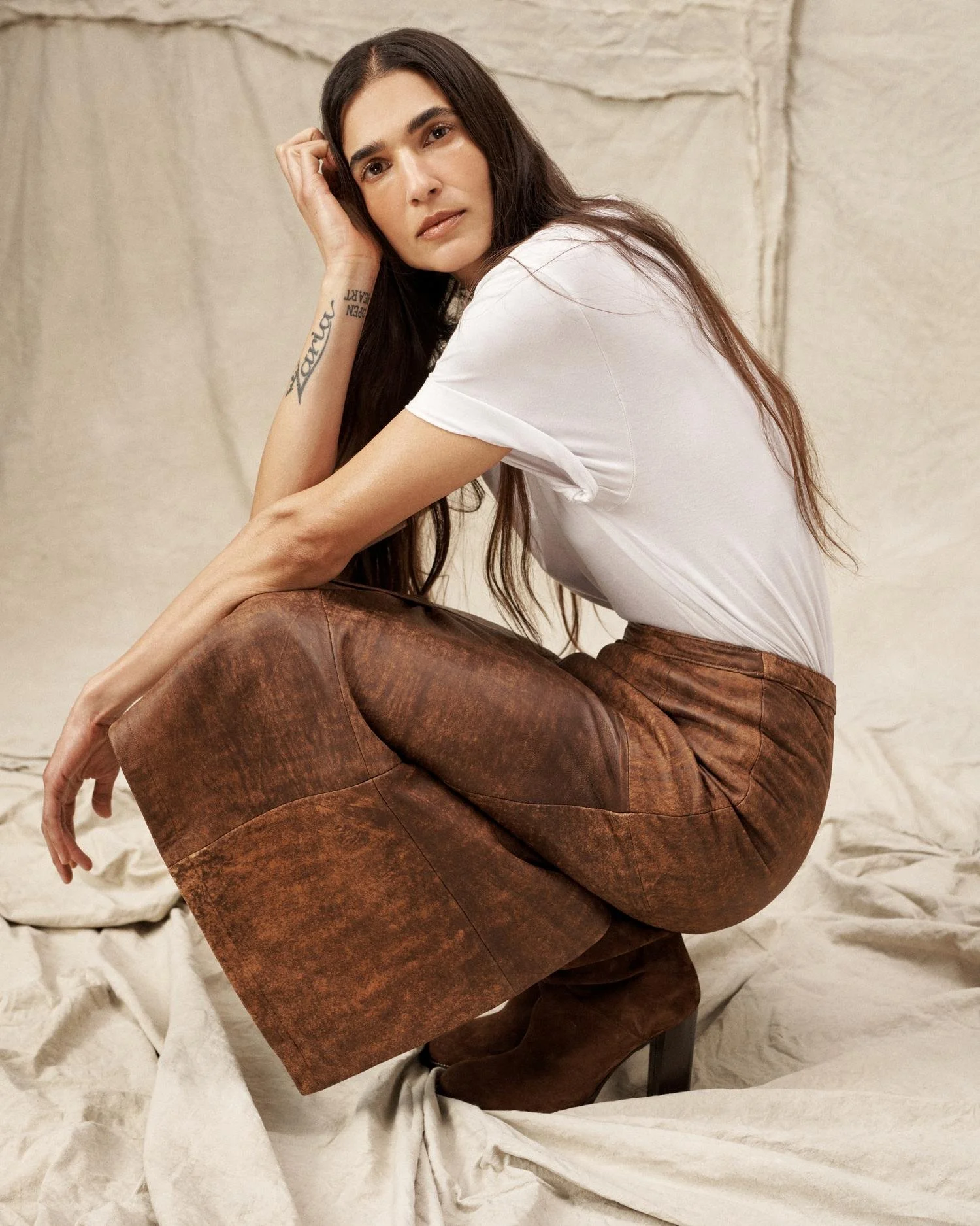 A woman with long dark hair sitting on the ground, wearing a white t-shirt, brown leather pants, and high heel boots. She has a tattoo on her left forearm and is looking into the camera with a neutral expression. The background is beige fabric.