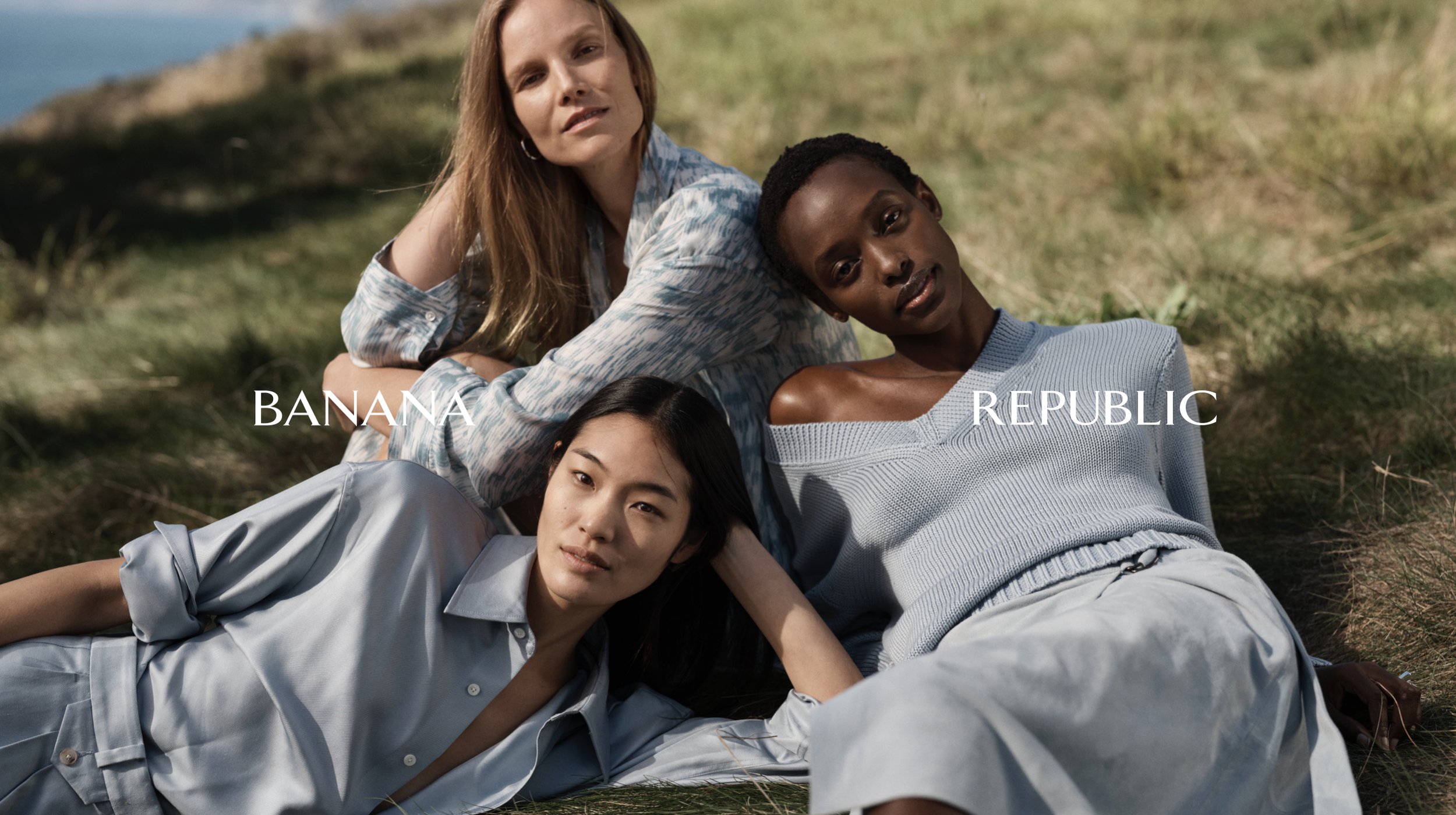 Four women lying on grass outdoors, with one woman resting her head on another's lap, and two women sitting behind them. The scene is sunny with a background of grass and a hill.