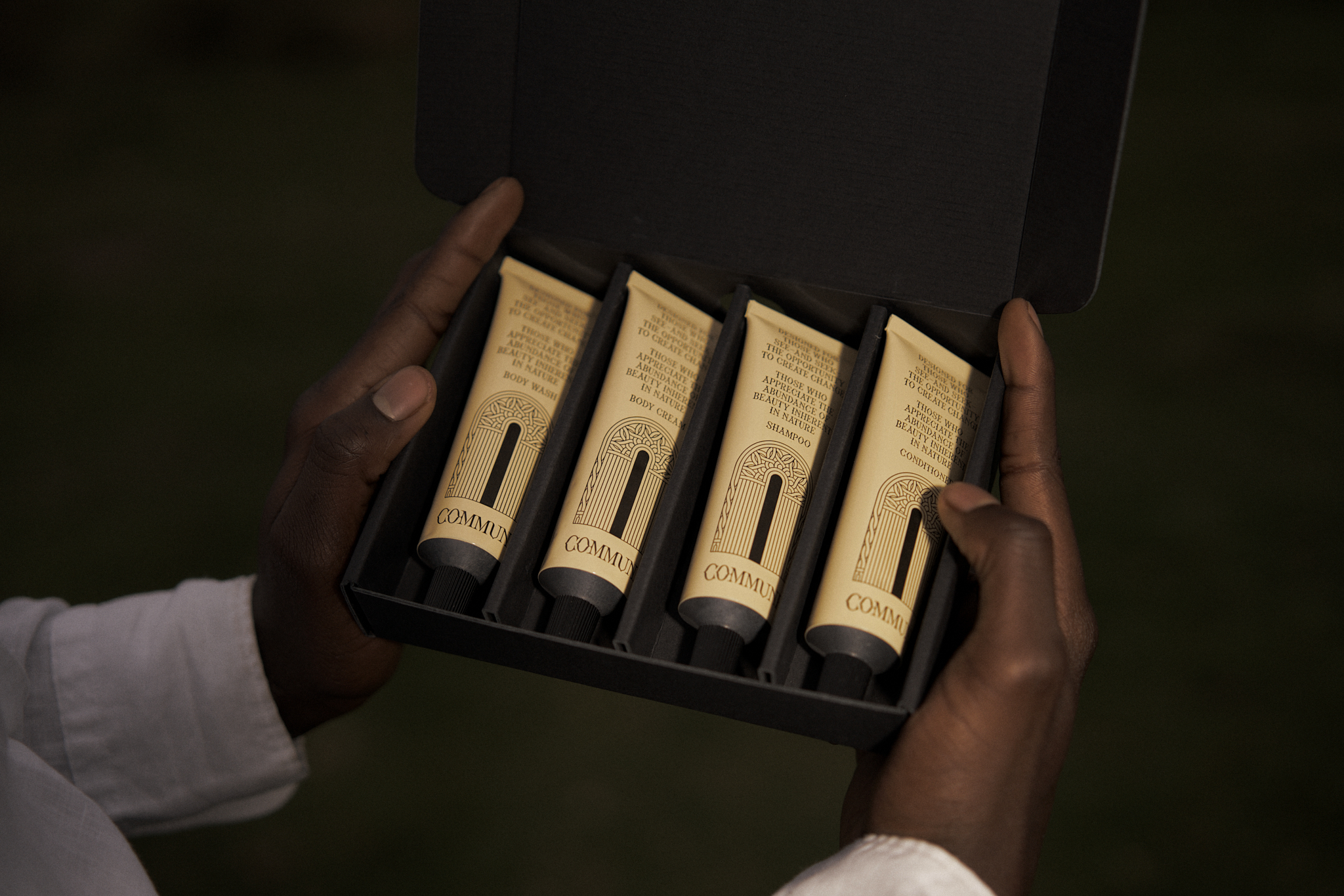 A person holding an open black box containing four tubes of skincare products labeled 'COMMUNITY' with decorative arch designs on the labels.
