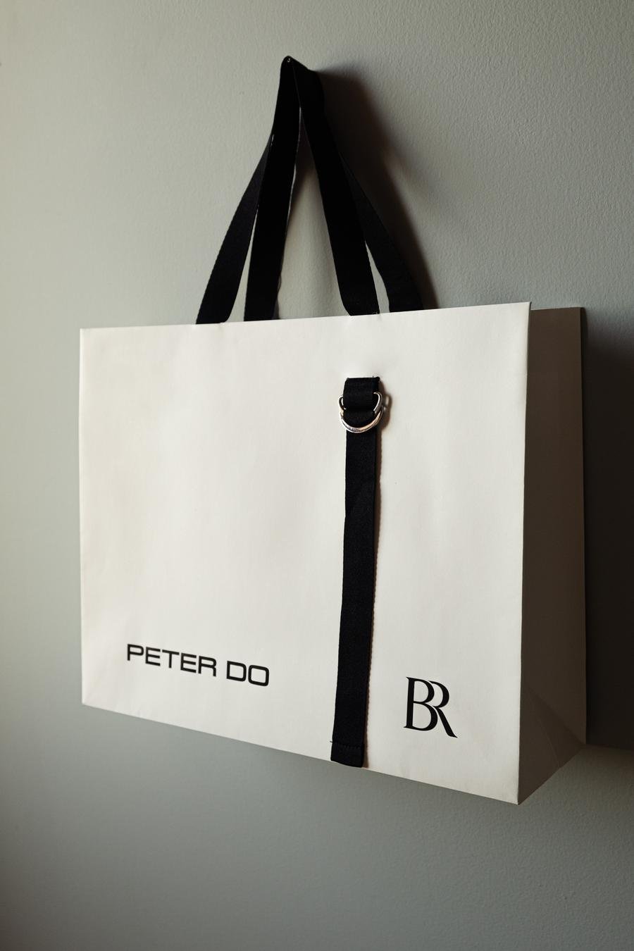 White Peter Do shopping bag with black handles hanging on the wall, featuring a black strap with a metal ring and the brand's logo.