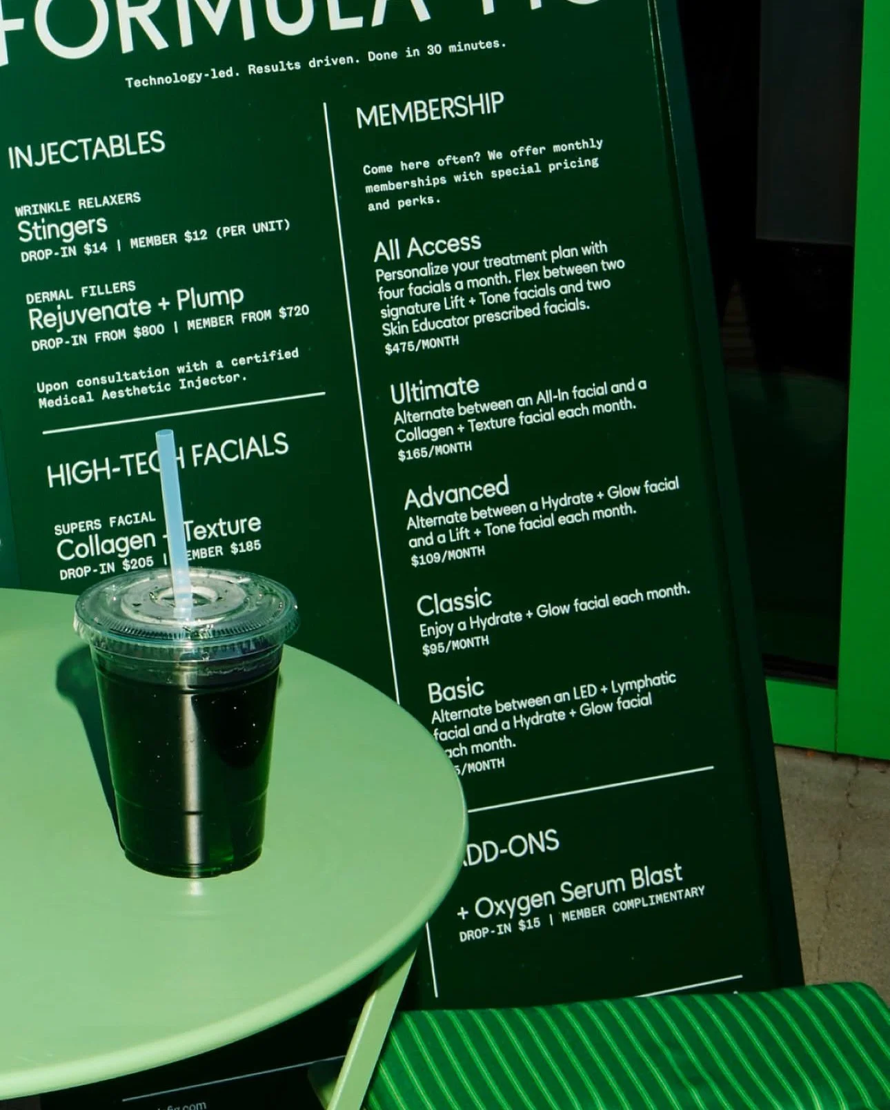 Close-up of a menu board for a facial treatment spa, with a dark beverage with a straw on a green table in the foreground.