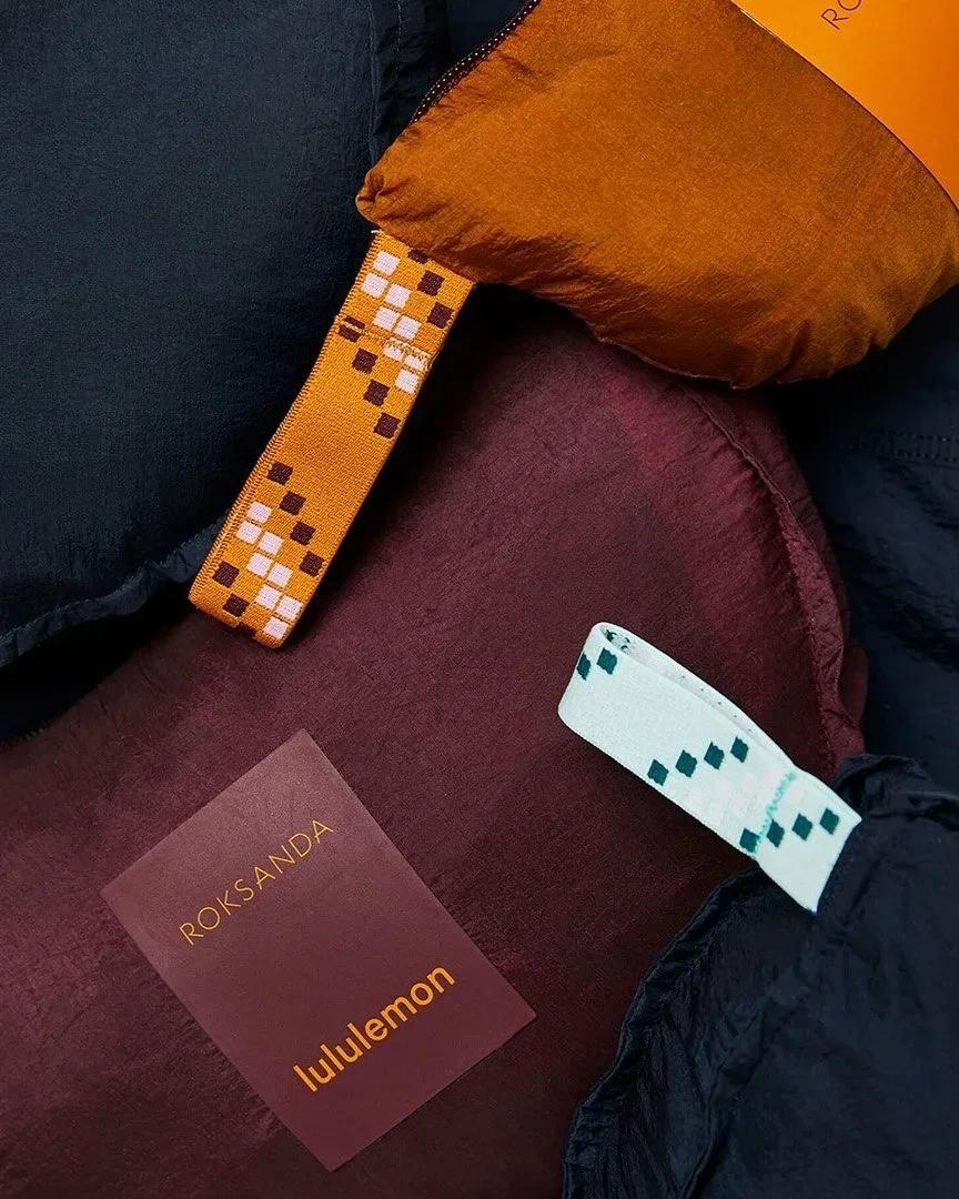 A close-up of two sleeping bags, one orange and one dark red, with digital patterned straps. There is a maroon card with yellow text reading 'ROKSANDA' and 'lululemon' placed on the fabric. The background includes dark fabric and a black material.