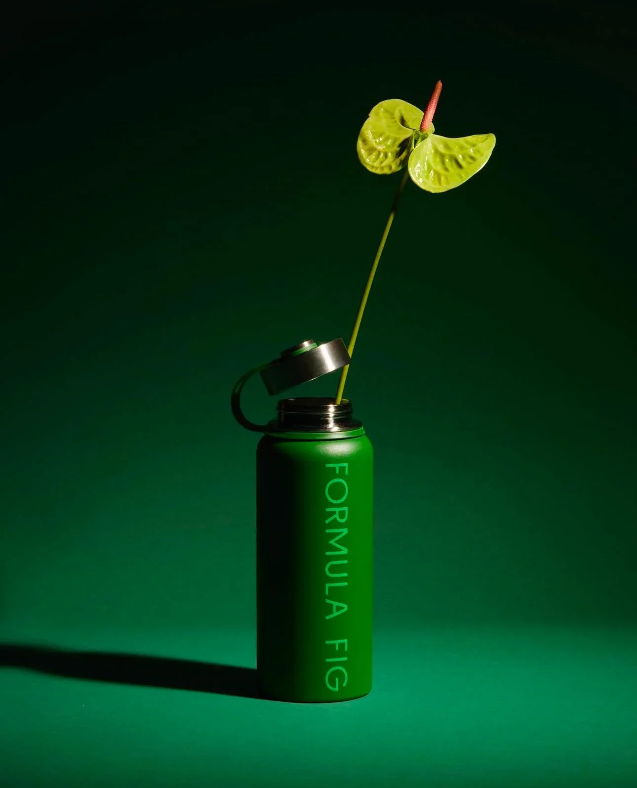 A green cylindrical container with the lid partially open, revealing a plant stem with two large green leaves in a vaporizer or spray can, set against a dark green background.