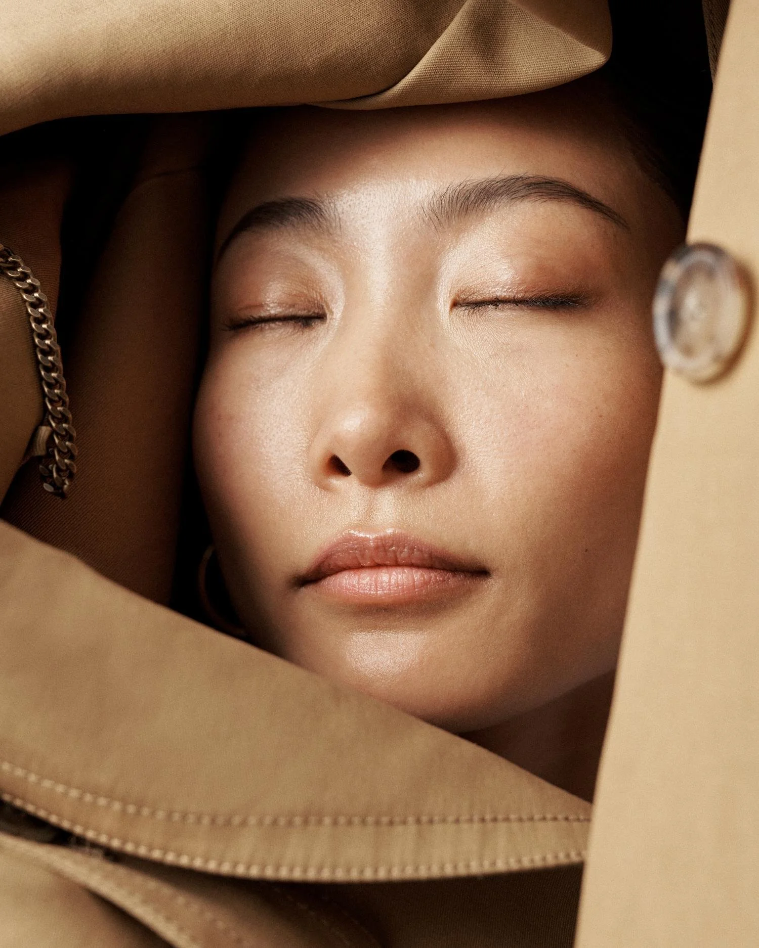 Close-up of a woman with closed eyes, wearing a beige jacket, a chain bracelet, and gold hoop earrings, with a neutral background.