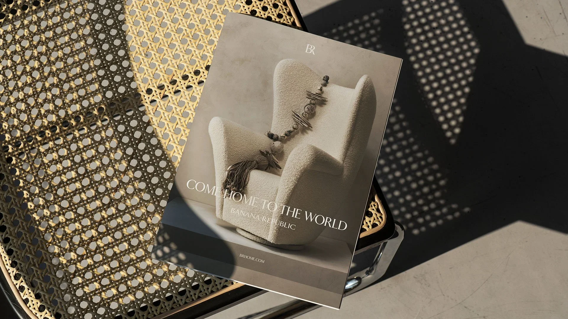 Magazine titled 'Come Home to the World' featuring a plush armchair with a beaded necklace on it, placed on a cane and metal framed chair, with shadows of a patterned object on the floor.