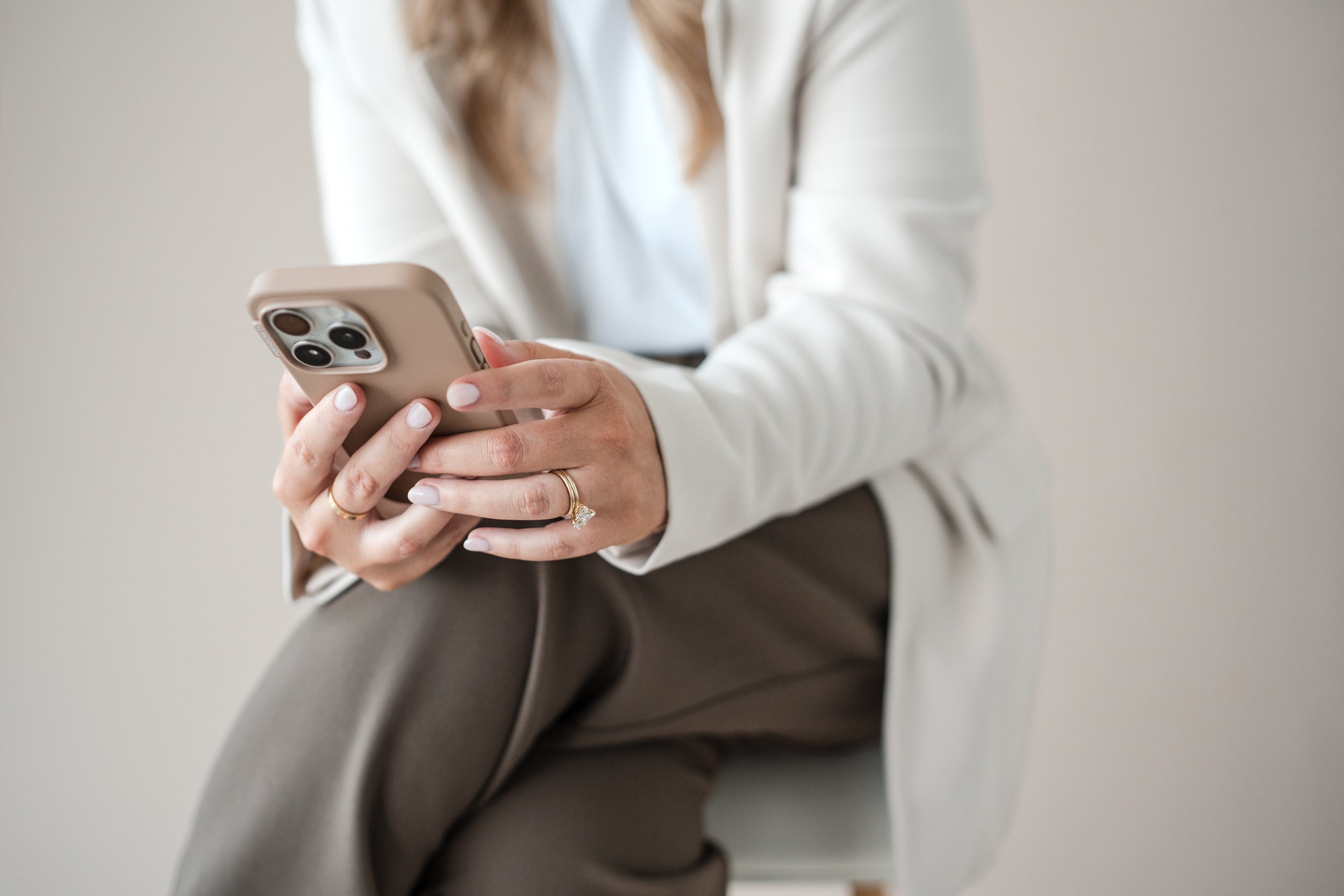 Person holding a smartphone with both hands, wearing a ring on one finger, dressed in a white top and brown pants, sitting on a chair against a plain background.
