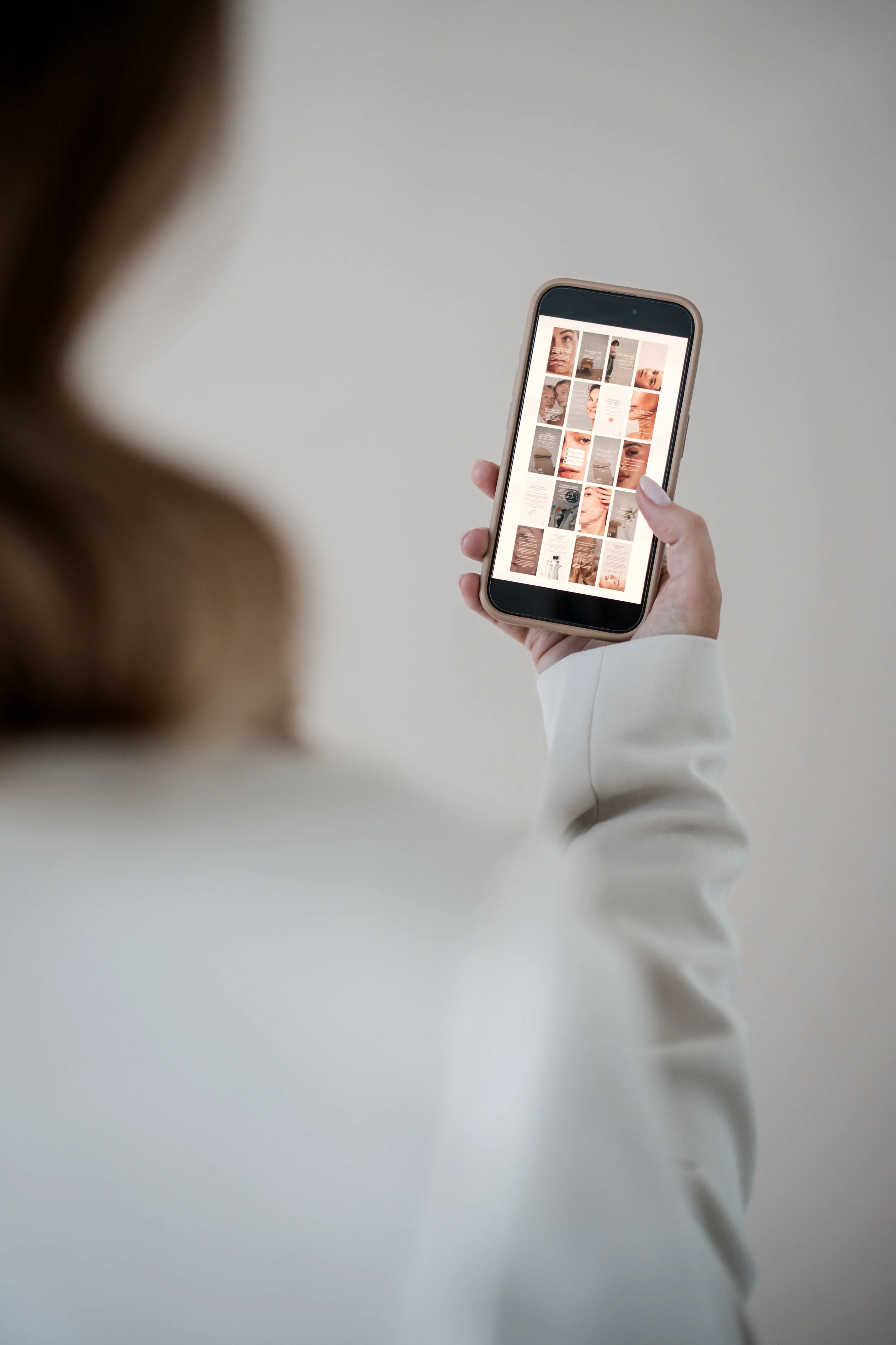 Person holding a mobile phone displaying a grid of images, mostly close-ups of faces and beauty products, against a plain, light-colored background.