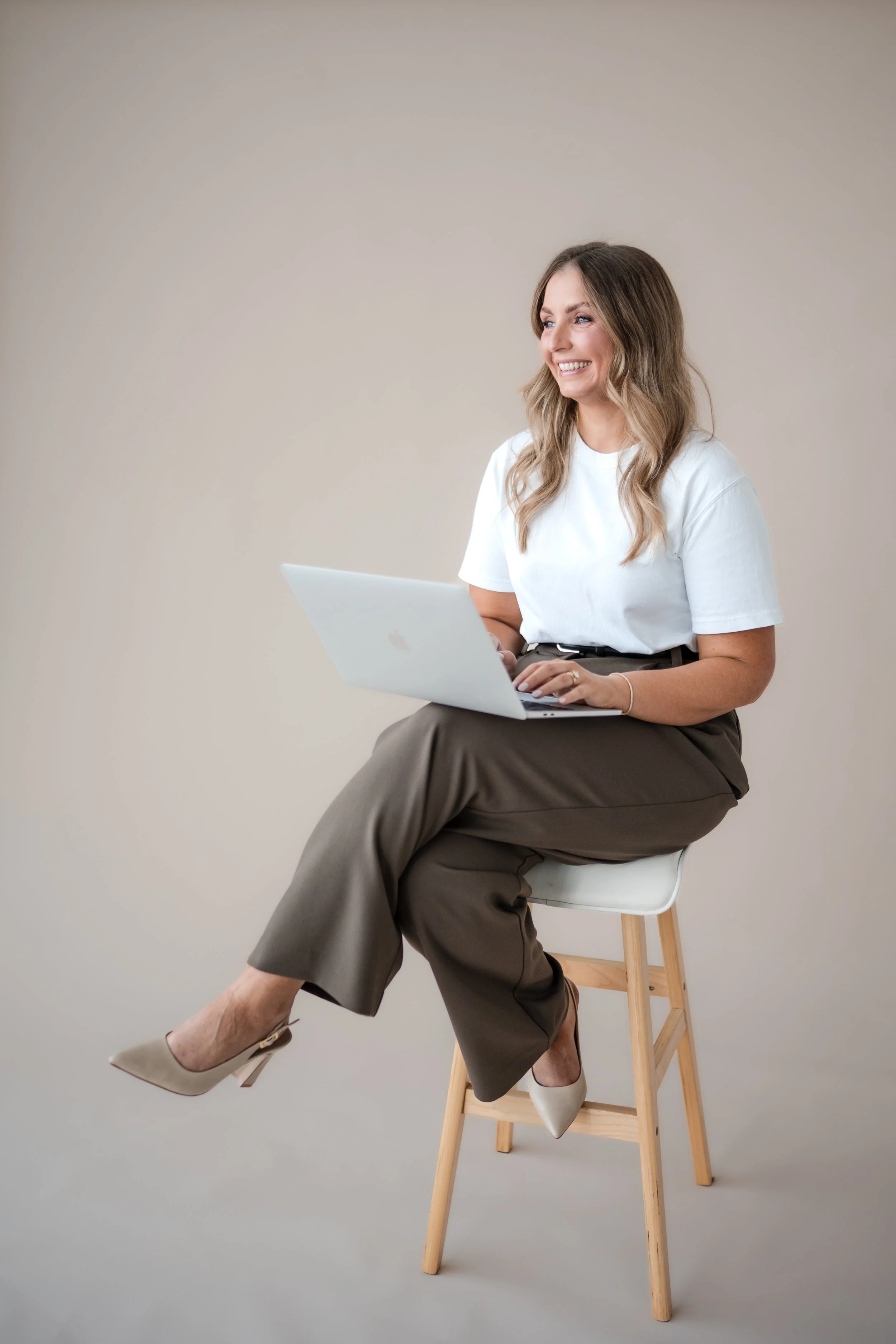 A woman with wavy, shoulder-length hair sitting on a wooden stool against a plain, light-colored wall. She is smiling and looking to her right while using a silver laptop. She is dressed in a white t-shirt, gray high-waisted trousers, and beige high heels.