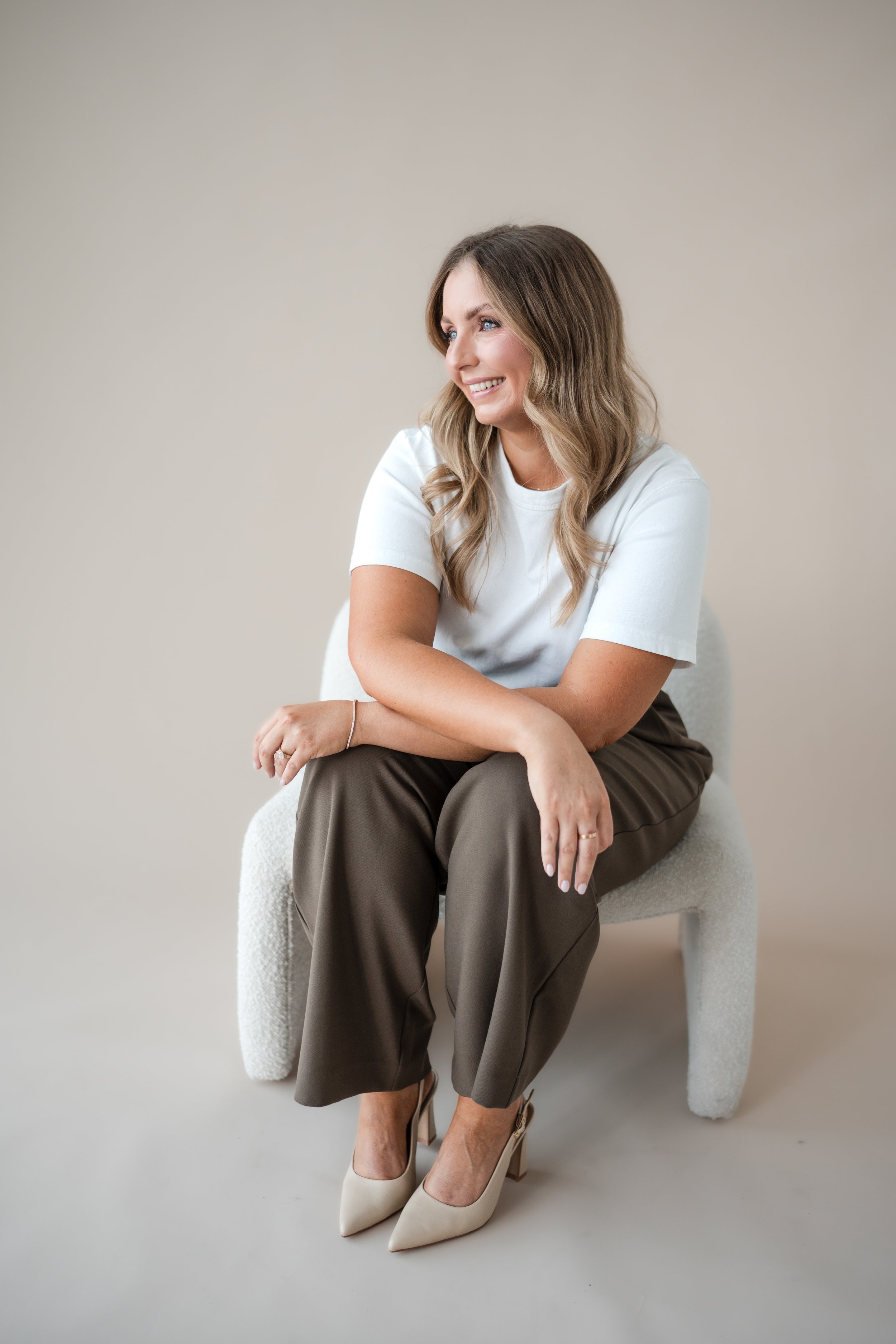 A woman with long wavy blonde hair sitting on a white textured chair against a plain neutral wall, wearing a white t-shirt, brown high-waisted pants, and cream-colored high heels, smiling and looking to her right.