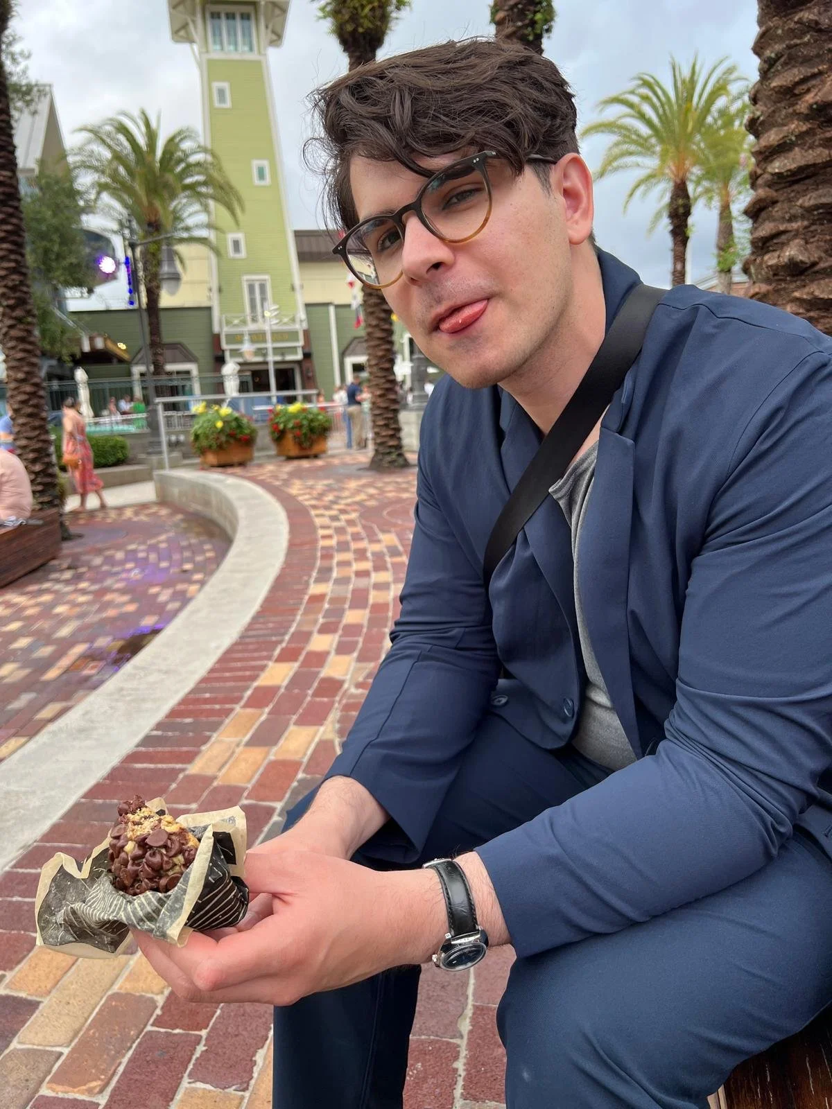 A young man with glasses and a watch, wearing a blue jacket, is sitting outdoors on a brick-paved area near palm trees, holding a chocolate dessert with toppings. He is sticking out his tongue playfully.