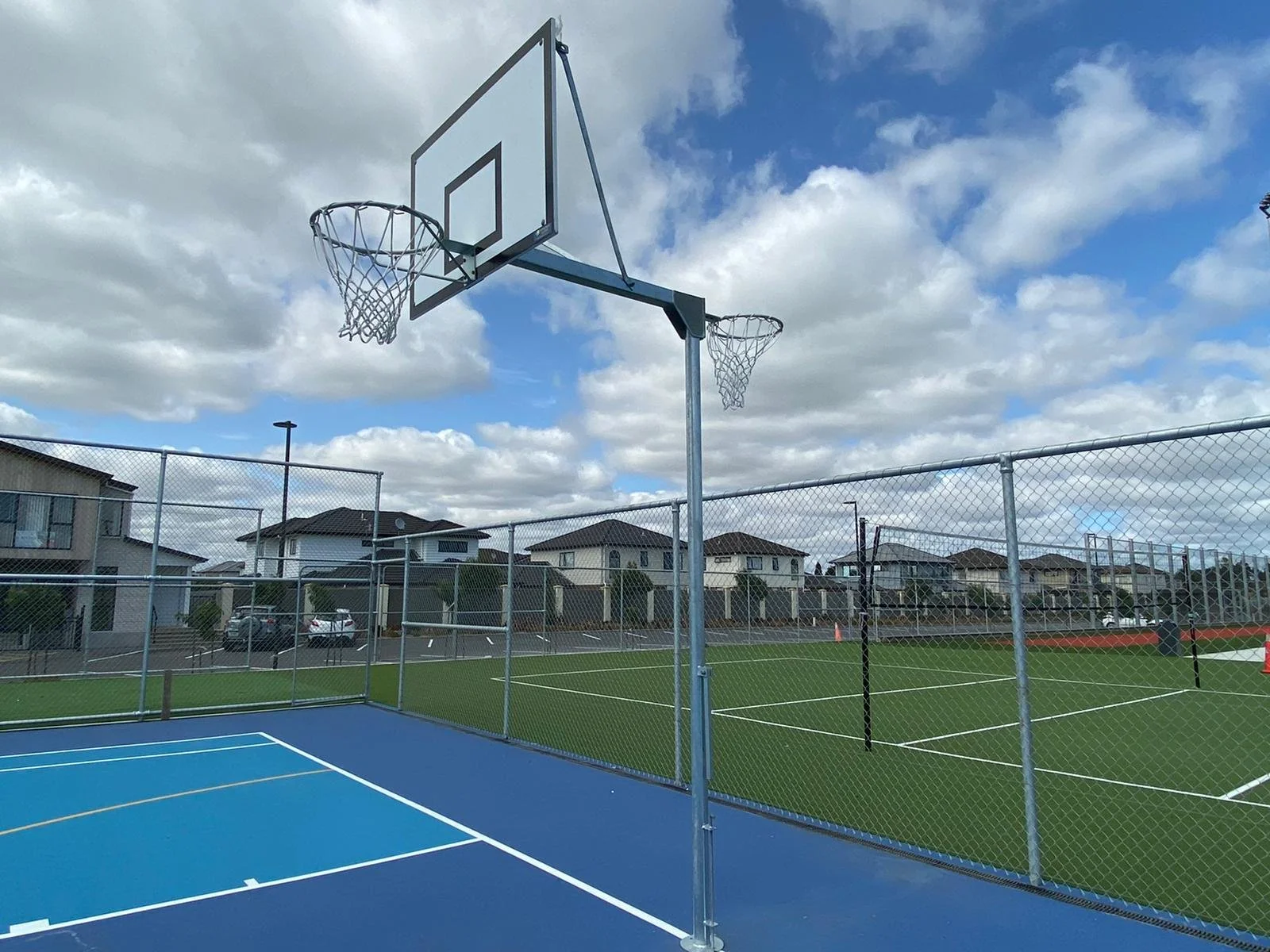 An outdoor sports court with a blue basketball court and tennis courts, surrounded by a chain-link fence, with residential houses and parked cars in the background under a cloudy sky.