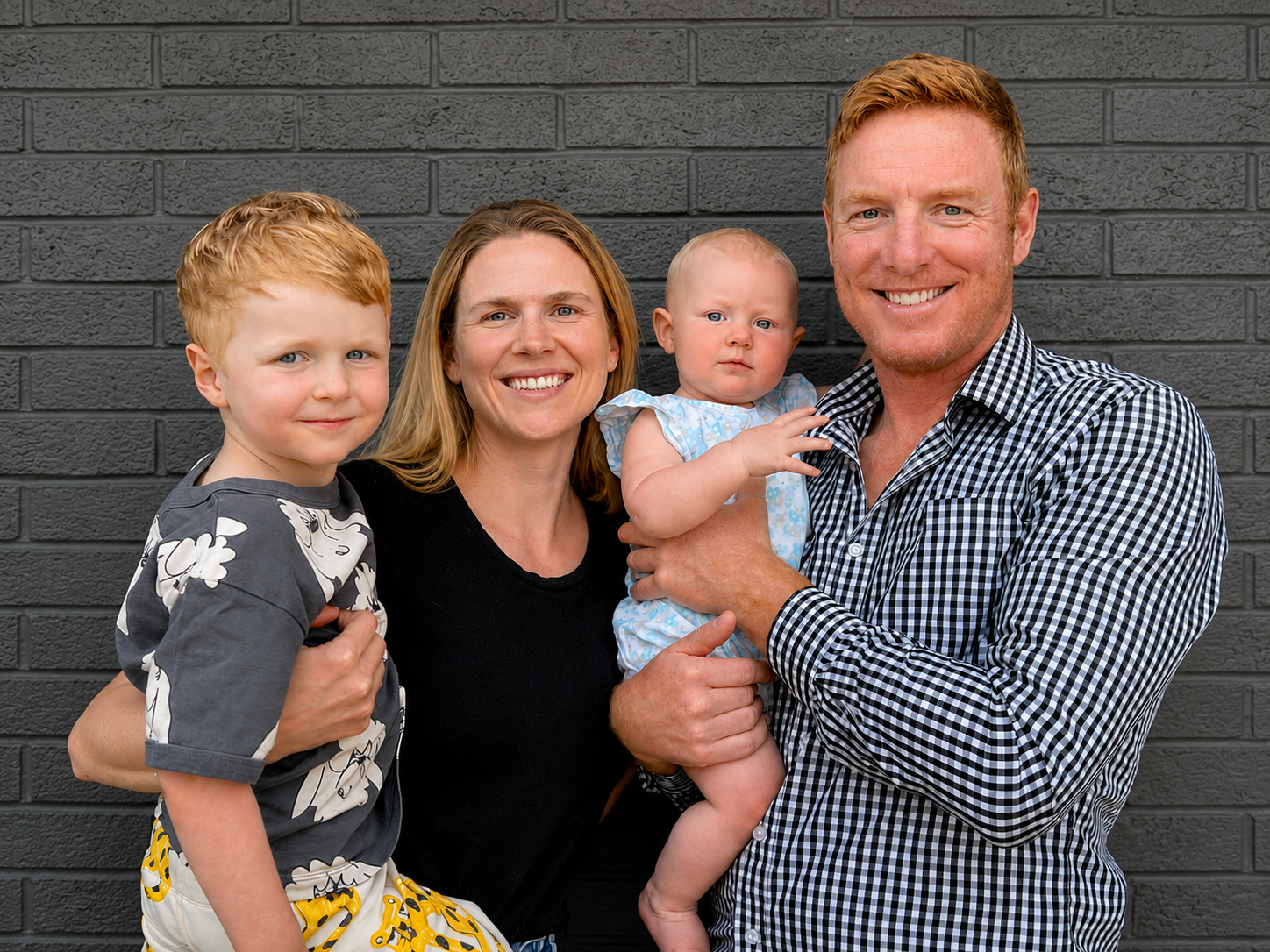 A family of four standing in front of a grey brick wall, smiling. The family includes a young boy with red hair wearing a patterned shirt, a woman with blonde hair in a black shirt, a baby girl in a light blue dress, and a man with red hair and a checkered shirt holding the baby.