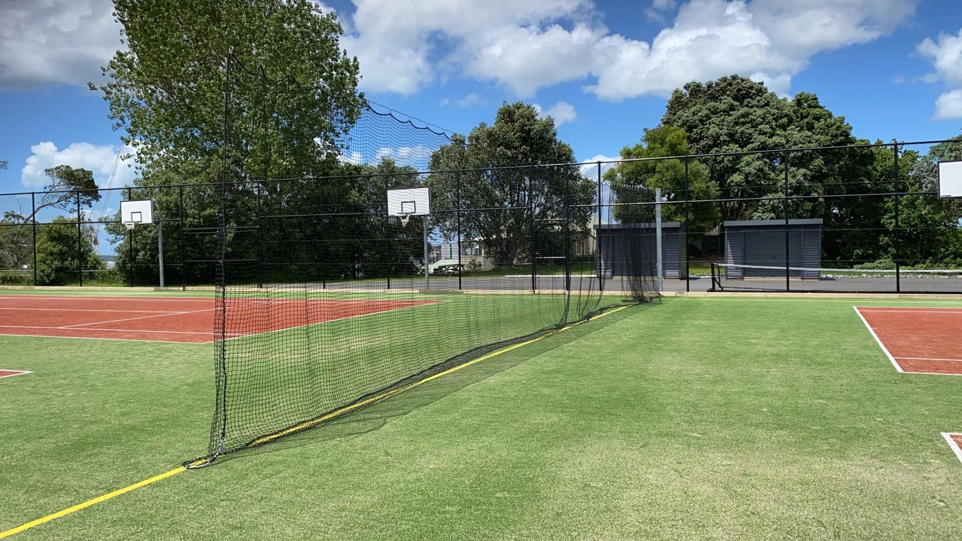 An outdoor pickleball or tennis court with red and green surfaces, surrounded by a tall black fence. There are two white basketball hoops and trees in the background, with a blue sky and some clouds.