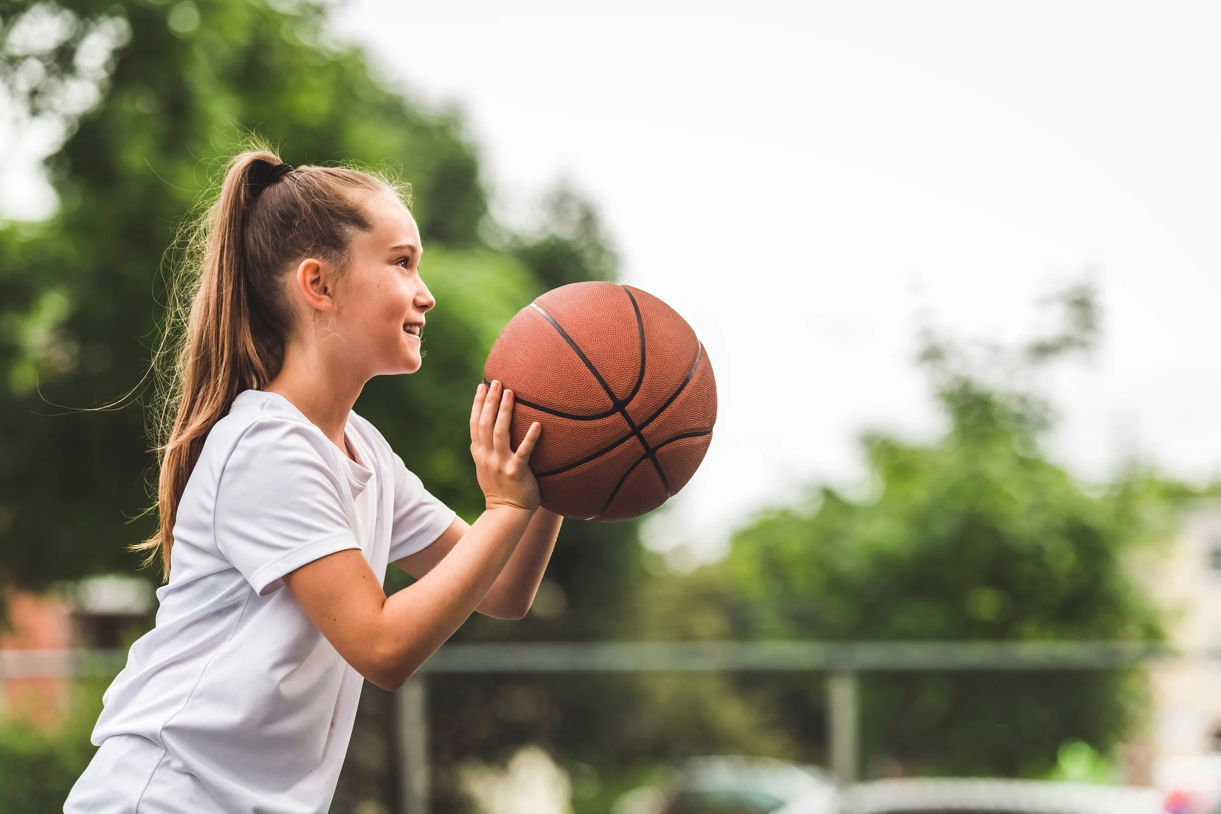 A young woman holding a basketball while outdoors, smiling, with green trees in the background.