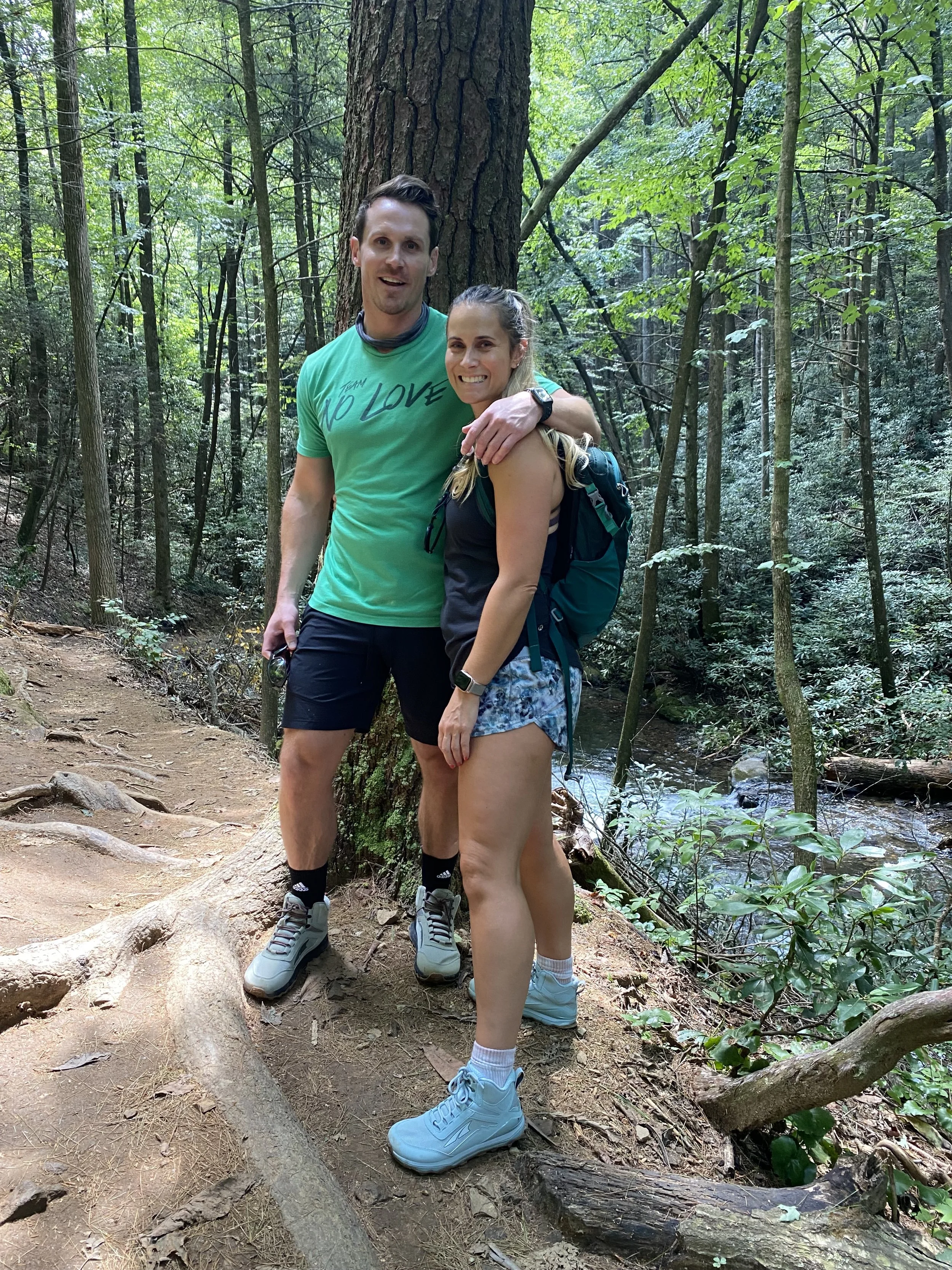 A man and a woman standing together on a forest trail near a stream, smiling and embracing, dressed in outdoor hiking gear.