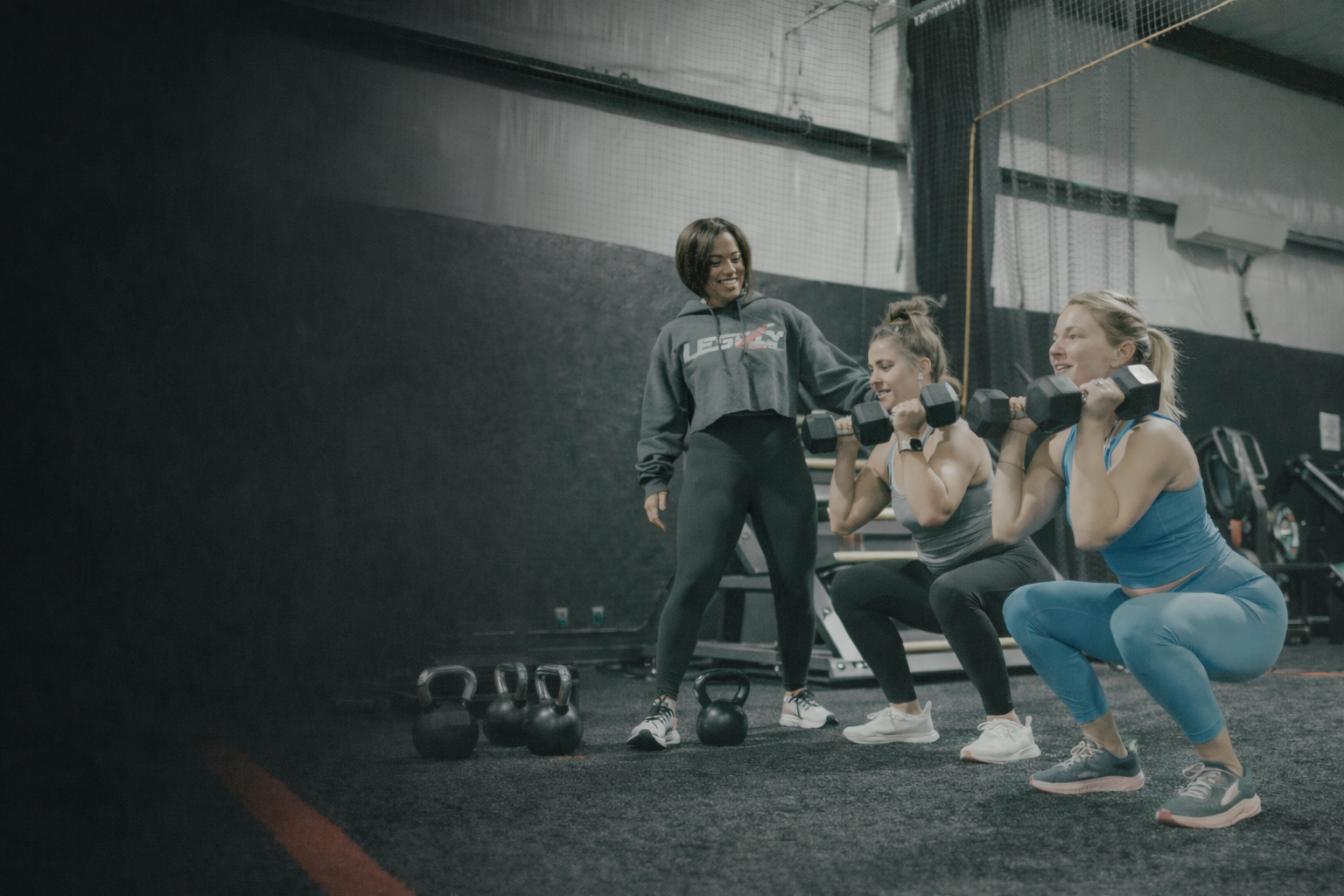 Three women lifting dumbbells during a workout session with a coach in a gym.