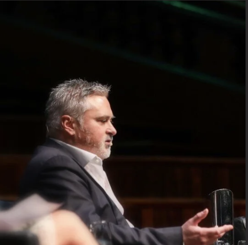 A middle-aged man with gray hair and a beard in a suit sitting at a table.