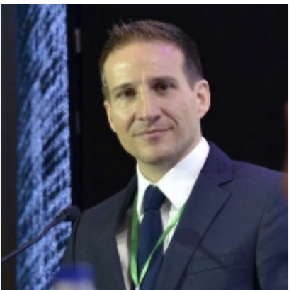 Leonidas Karagounis wearing a suit and tie, standing in front of a screen displaying data or text.