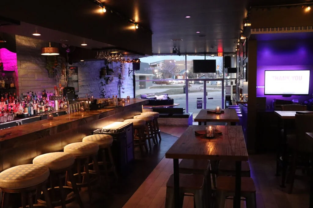 Interior of a cozy bar with a long wooden counter, barstools, and ambient lighting. There are tables with candles, a large window showing an outside street scene, and a lit 