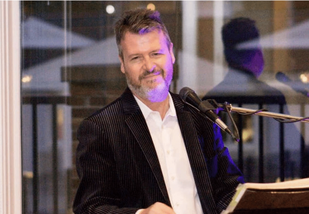 A man with a beard and brown hair, wearing a black pinstripe suit and white shirt, is smiling while standing at a microphone, with a reflection of himself in a glass window behind him.