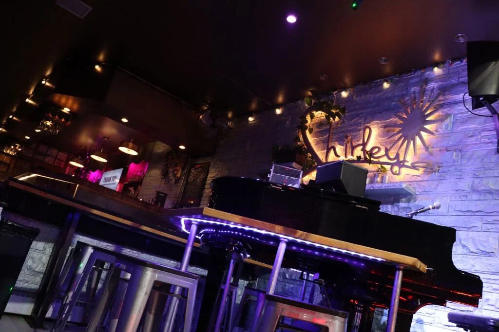 Nightclub stage with a grand piano, illuminated sign that says 'Hibiki', and a plant, with purple and yellow lights creating a vibrant atmosphere.