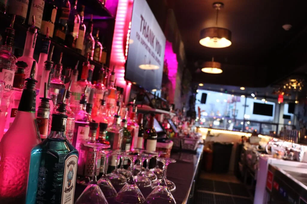 A bar with shelves stocked with bottles of liquor, pink lighting illuminating the bottles, and overturned cocktail glasses on the counter. The background shows a dimly lit lounge with tables, chairs, and large windows.