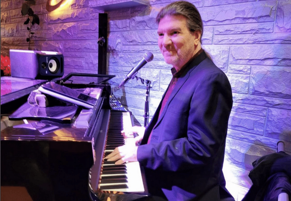 A man in a dark blazer playing a grand piano at a venue with purple lighting and a white brick wall backdrop.