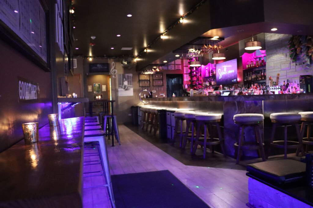 Interior of a dimly lit bar with purple lighting, a bar counter with stools, shelves with bottles, and a TV screen, creating a cozy atmosphere.
