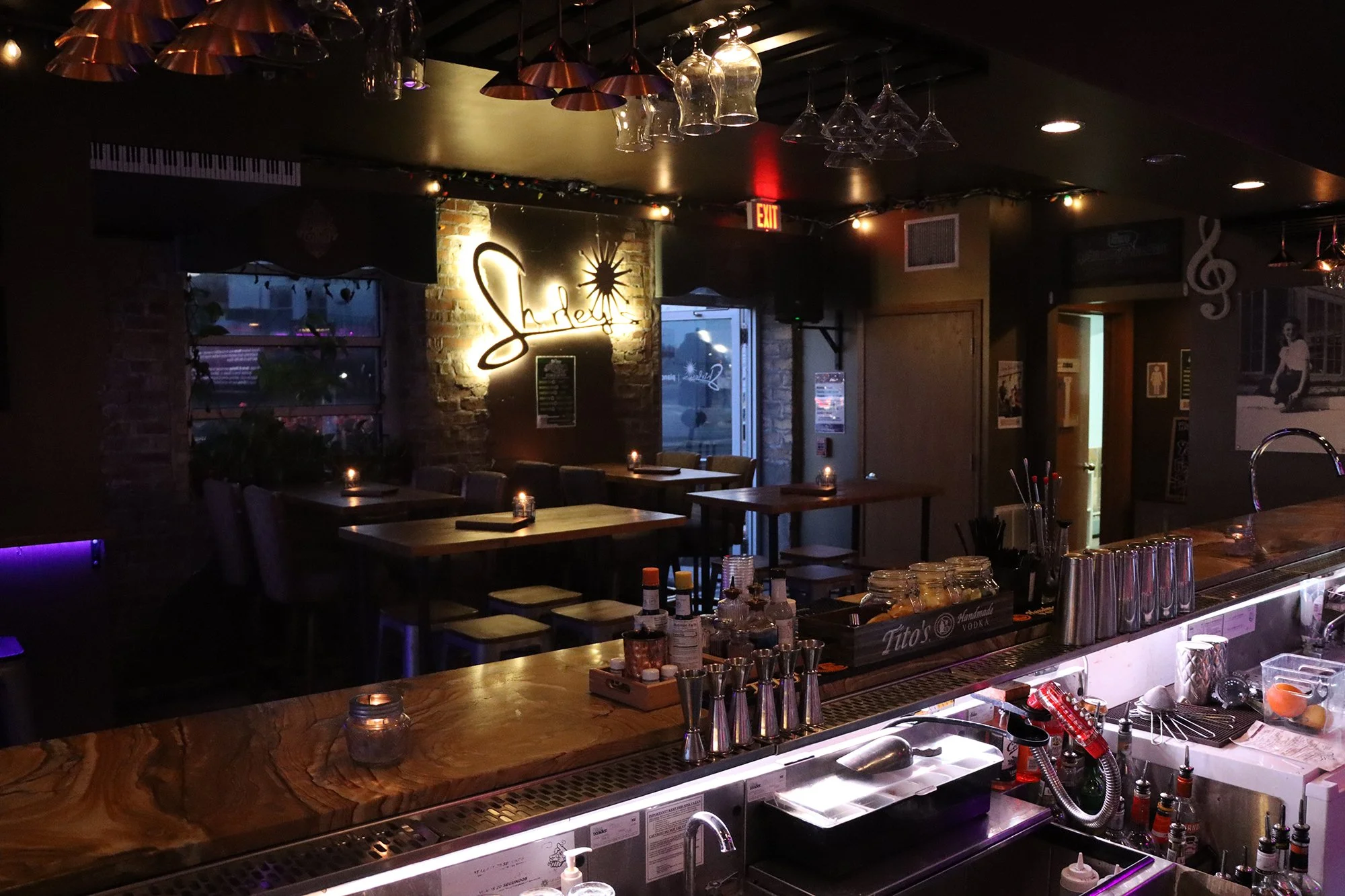 A dimly lit bar and restaurant interior with tables, chairs, and a brick wall. There is a neon sign on the wall that says 'Slice' with a starburst design. The counter has various bottles, jars, and bar tools. Glasses hang upside down from the ceiling