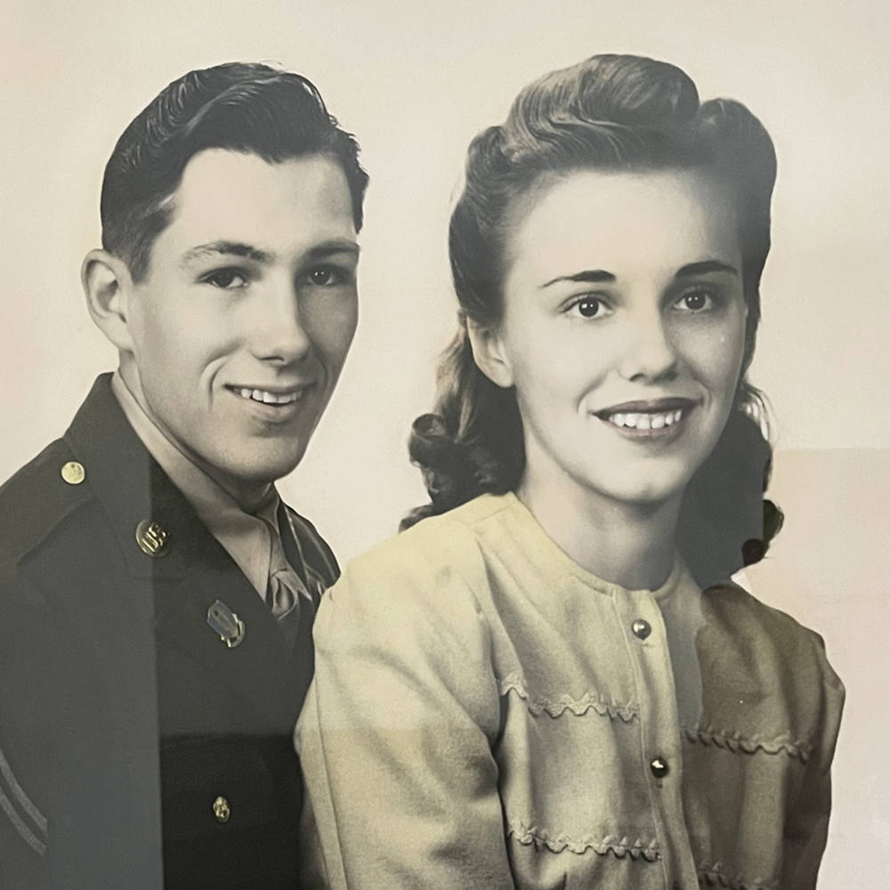A black and white photo of a young man in military uniform and a young woman in a light-colored dress, both smiling at the camera.