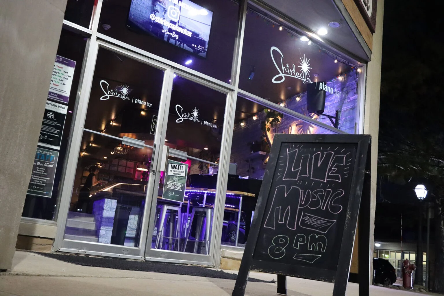 Exterior of a piano bar named 'Shirelle's' with a chalkboard sign announcing live music at 8 PM, nighttime scene, illuminated interior visible through glass doors.