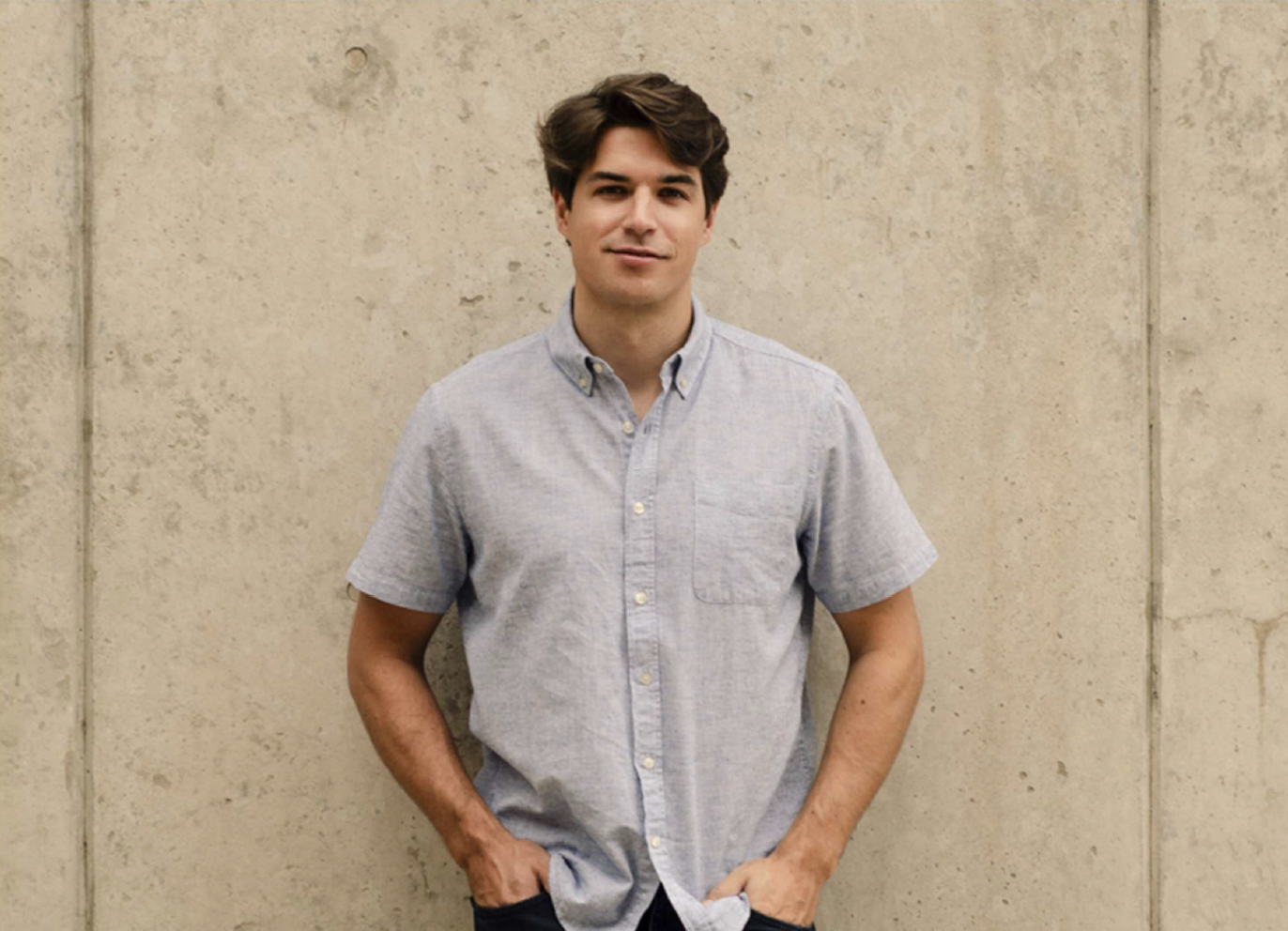 A young man with brown hair and fair skin standing against a beige concrete wall, wearing a light gray short-sleeve button-up shirt with his hands in his pockets.