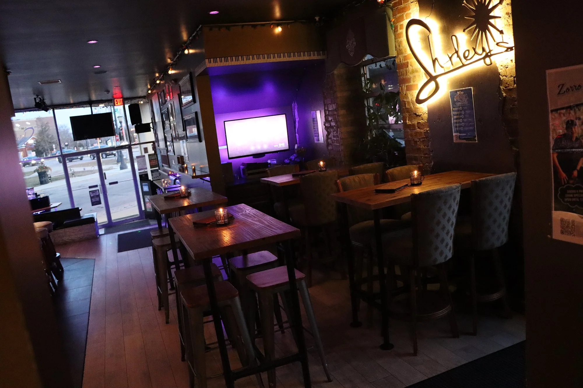 Inside a cozy bar or lounge with wooden tables, candles, a large neon sign that says 'Shelby', a TV mounted on the wall, and a view of the street through the glass front door.