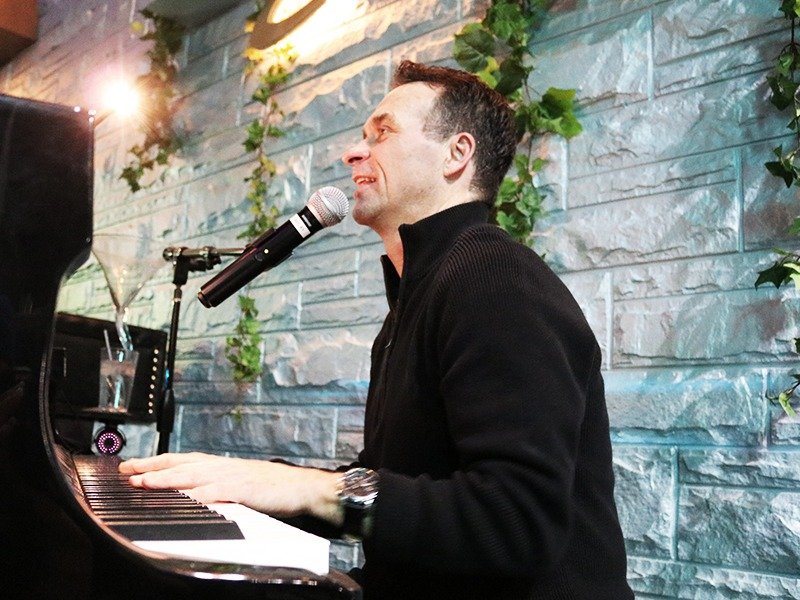 A man playing a grand piano and singing into a microphone in a decorated indoor setting with green plants and a stone wall.