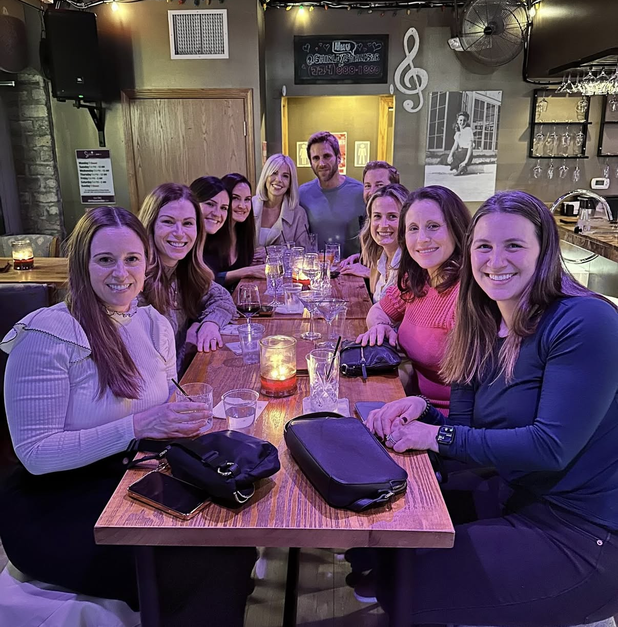 A group of eleven women and one man gathered around a wooden table in a restaurant, smiling for the camera. The table has drinks, candles, and personal belongings on it. The background features a chalkboard with music notes, a black-and-white photo of a girl, shelves with glasses, and a bar area.